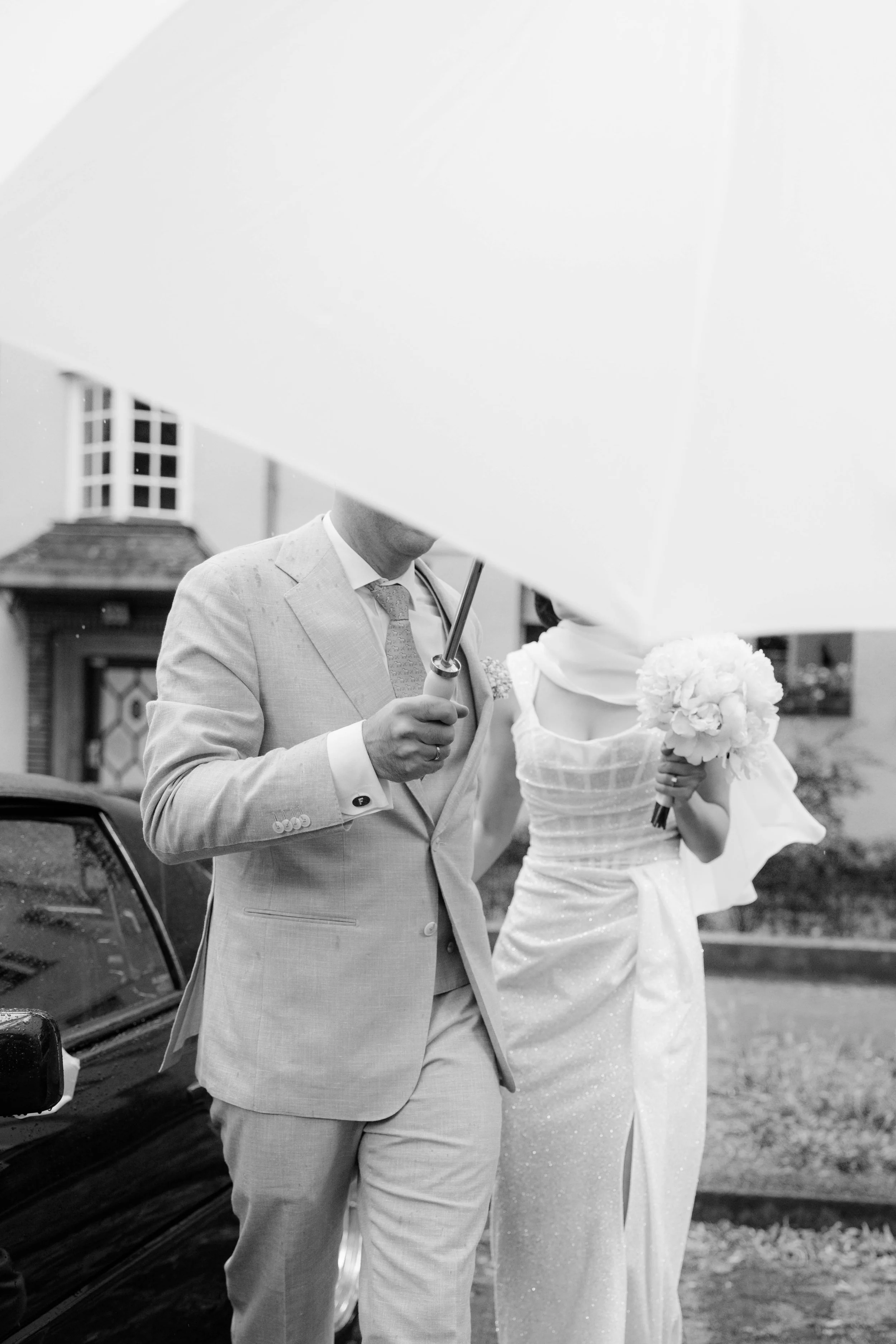 Ein Brautpaar steht im Regen unter einem großen Regenschirm, die Braut hält einen Blumenstrauß. Das Bild ist in Schwarz-Weiß.