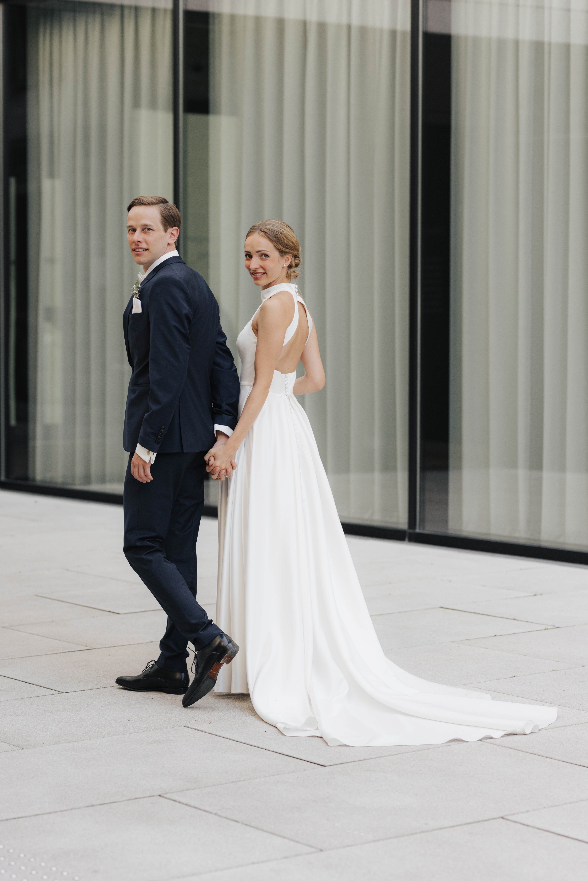 Ein Brautpaar hält Händchen, vor einem modernen Gebäude mit großen Glasfenstern.