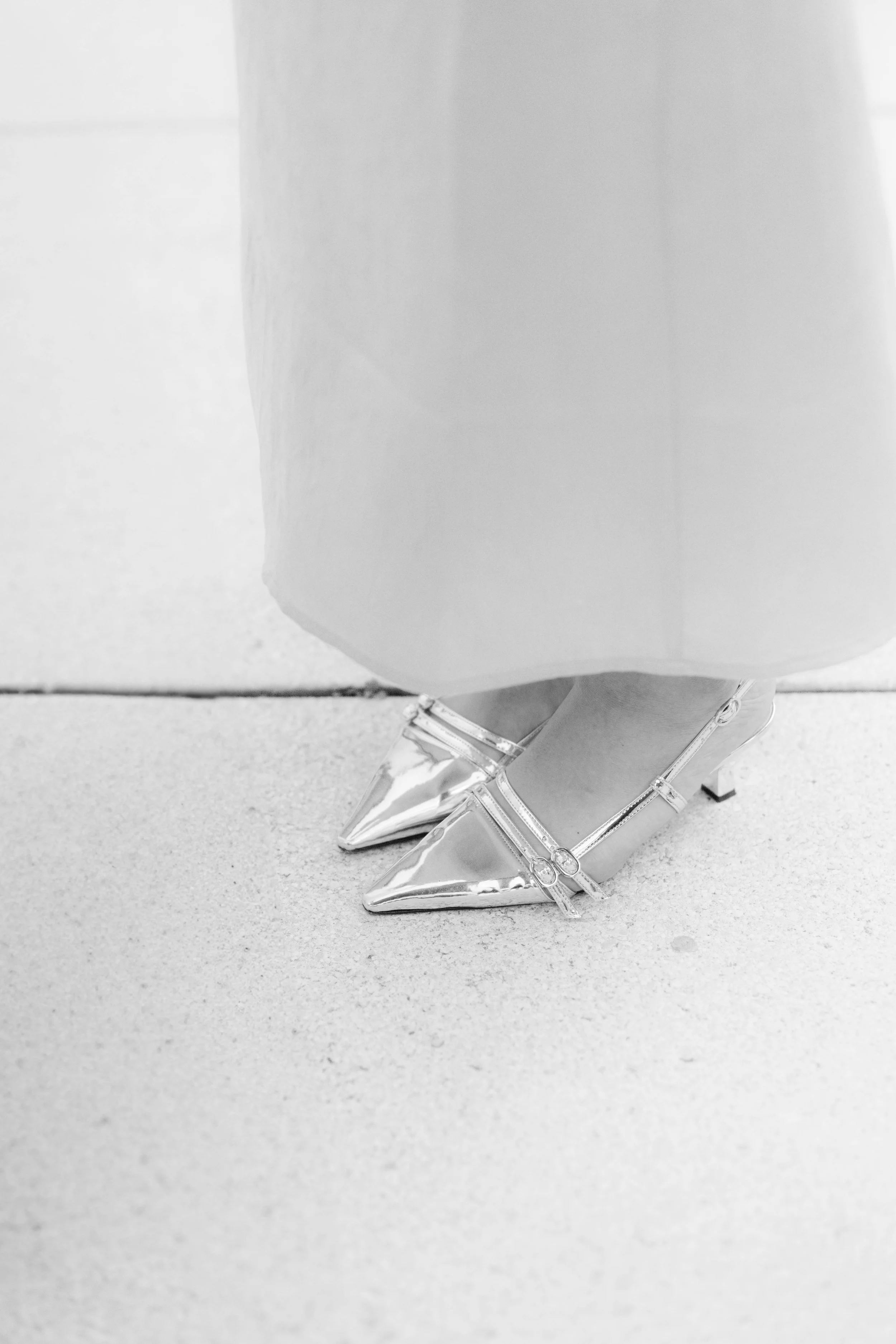 Nahaufnahme von silberfarbenen High Heels mit Riemen, getragen von einer Person in einem langen Kleid auf einer hellen Oberfläche.