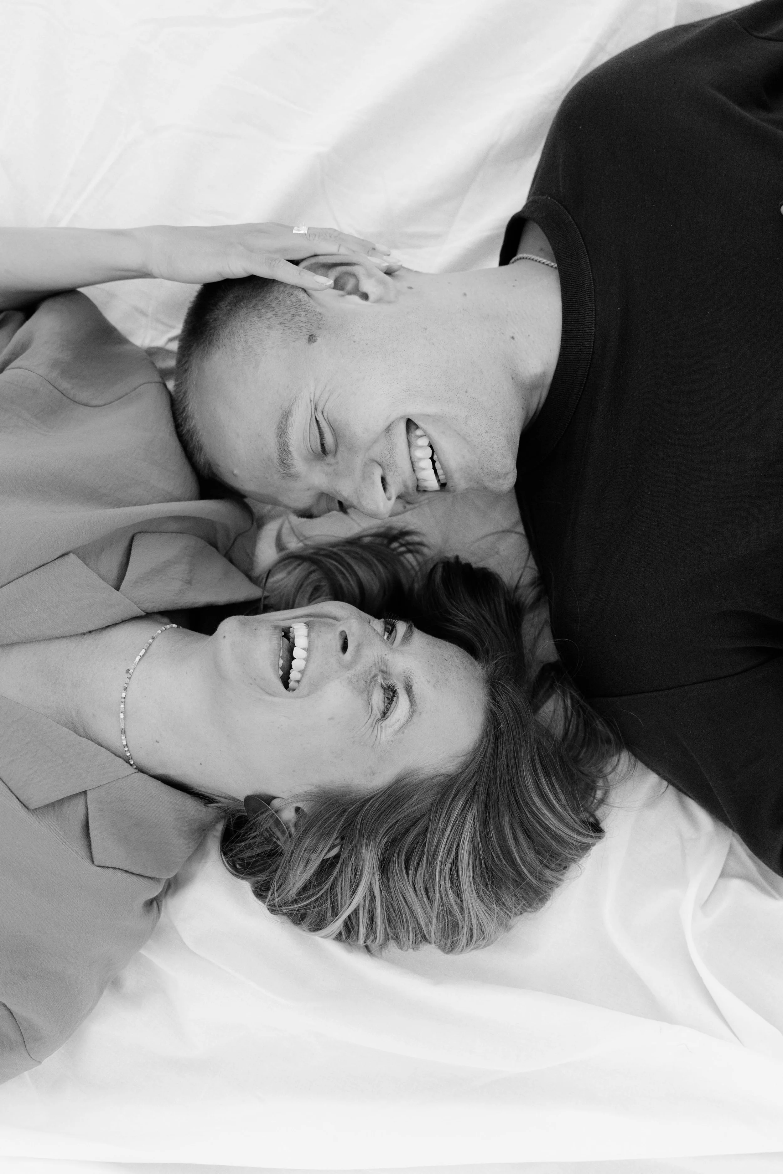 Ein Lächelndes Paar liegt auf einem Bett, die Frau berührt den Mann am Kopf, beide lachen und schauen sich fröhlich an.