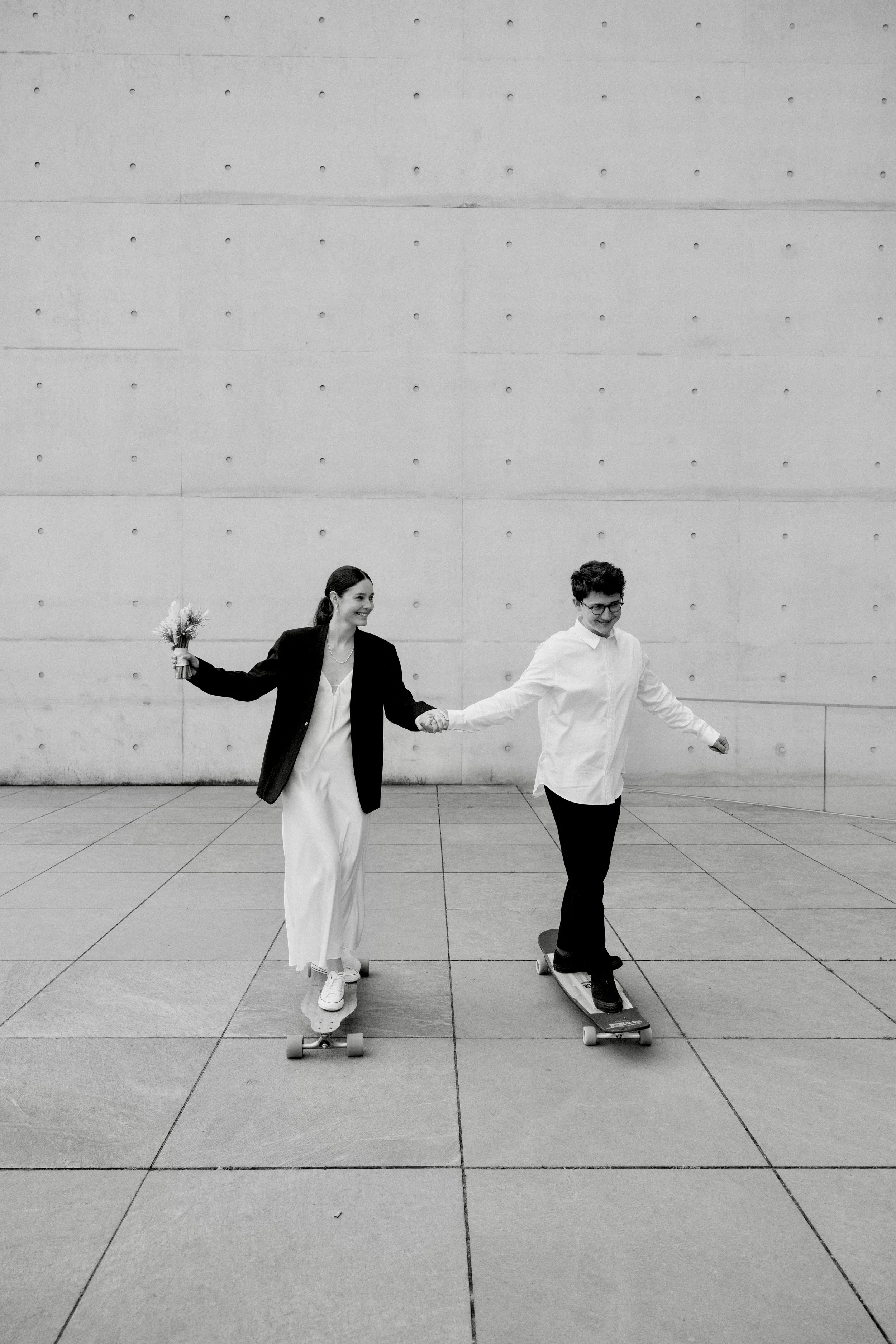 Ein Paar fährt auf Skateboards, die Frau hält einen Blumenstrauß und beide lächeln, gegen eine moderne Betonwand.