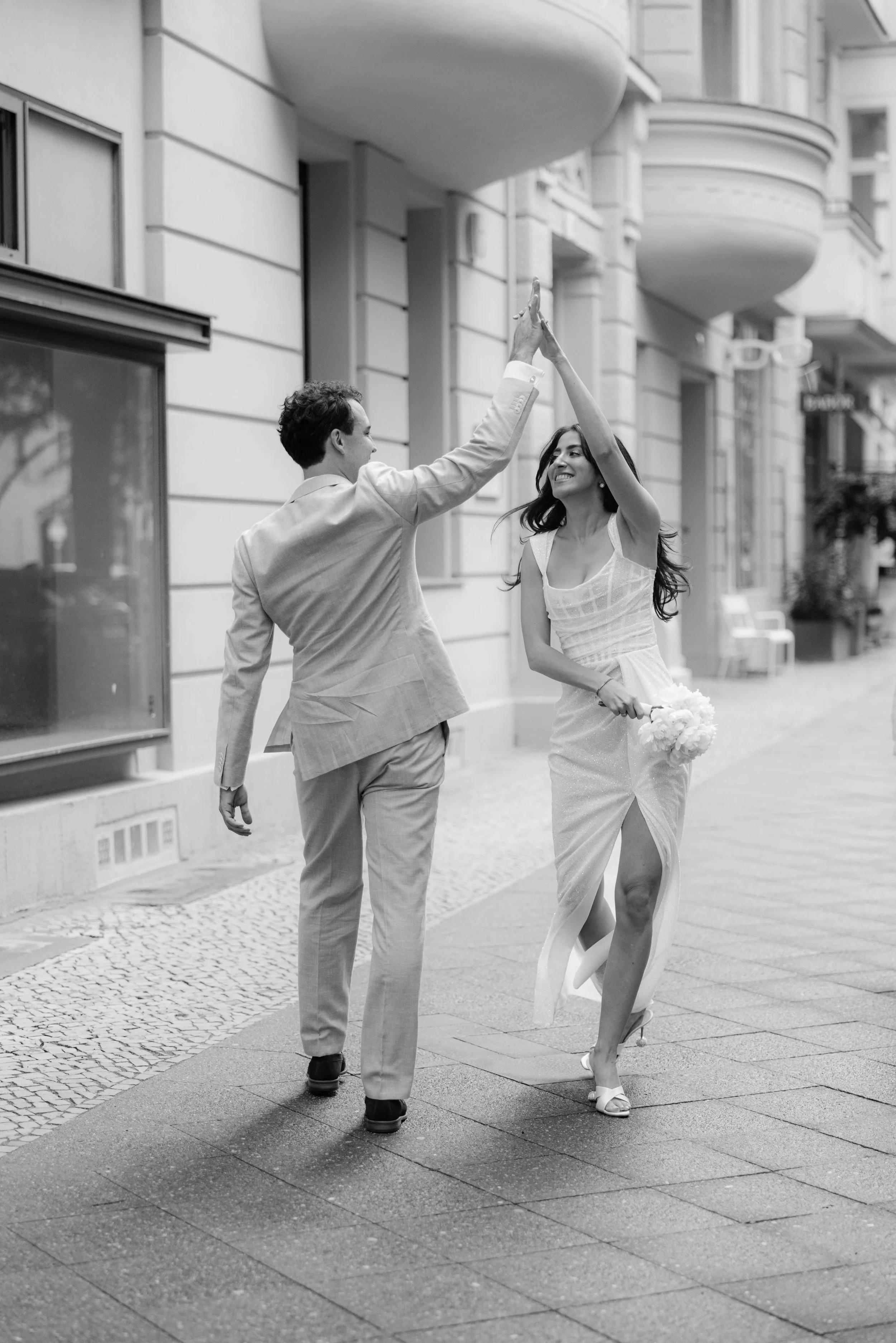 Ein glückliches Paar tanzt auf der Straße, die Frau trägt ein Hochzeitskleid und hält einen Blumenstrauß, während sie sich gegenseitig high-fiven.