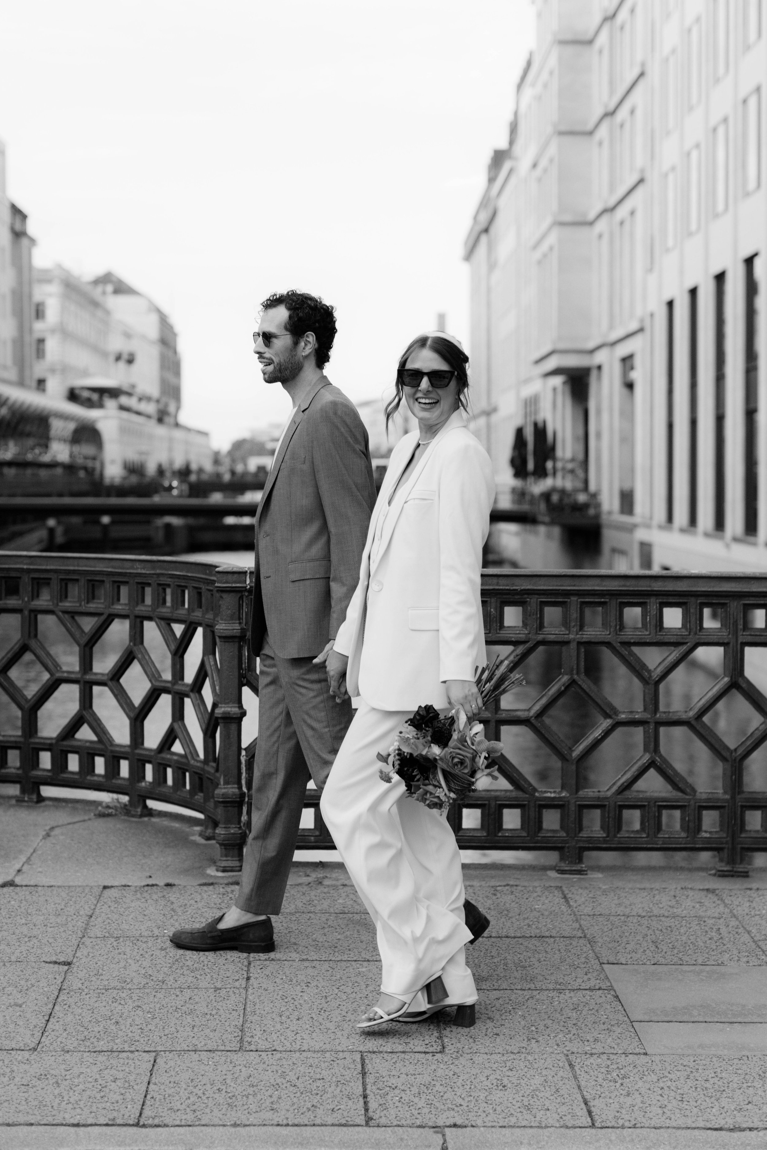 Schwarz-weiß-Foto eines lachenden Paares in Hochzeitskleidung, das auf einer Brücke in einer Stadt steht, Frau hält einen Blumenstrauß.
