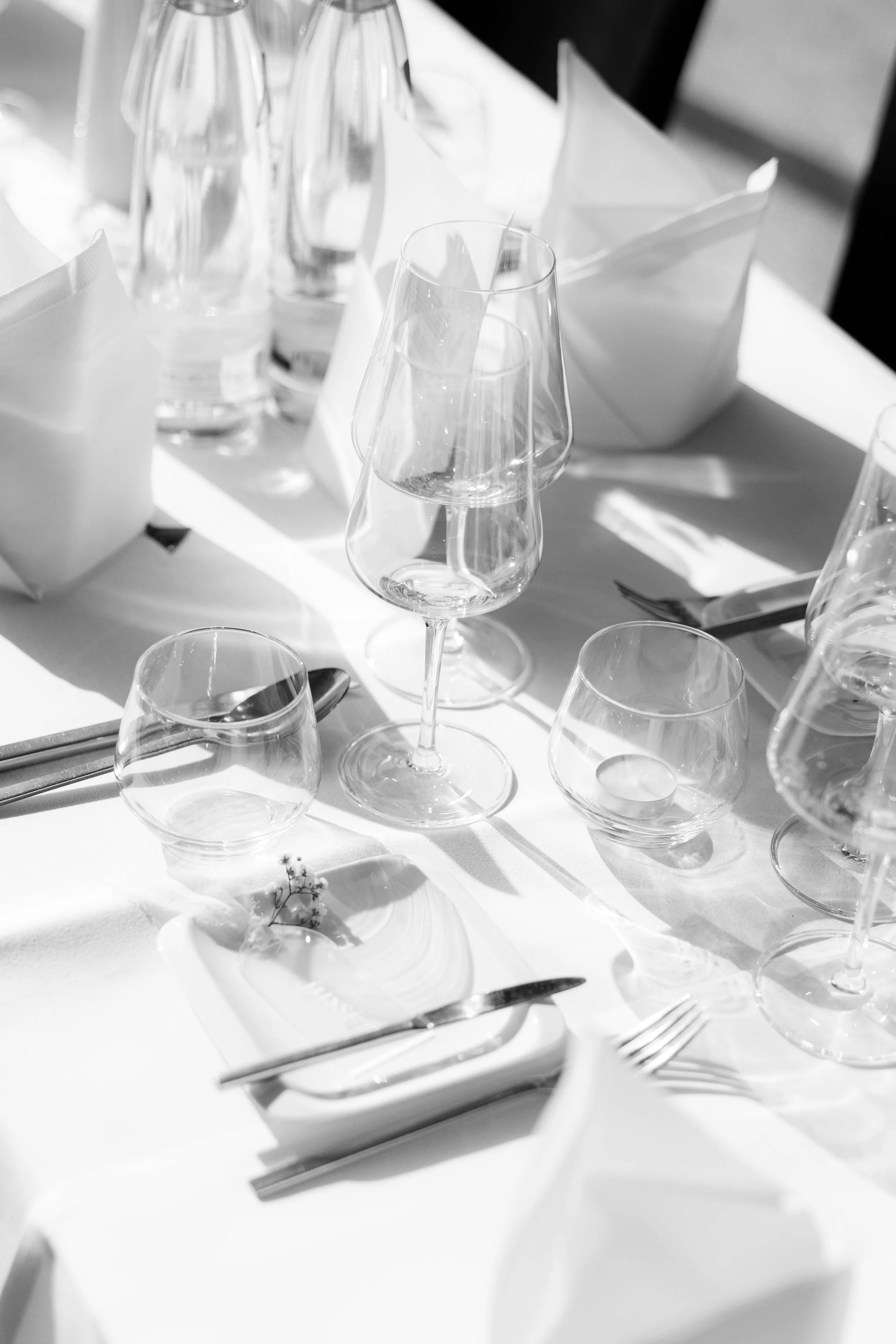 Weiß gedeckter Tisch mit Weingläsern, Wasserflaschen, Besteck, weißen Servietten und einer kleinen Dekoration. Blick auf einen elegant gedeckten Speisetisch bei einer Veranstaltung.