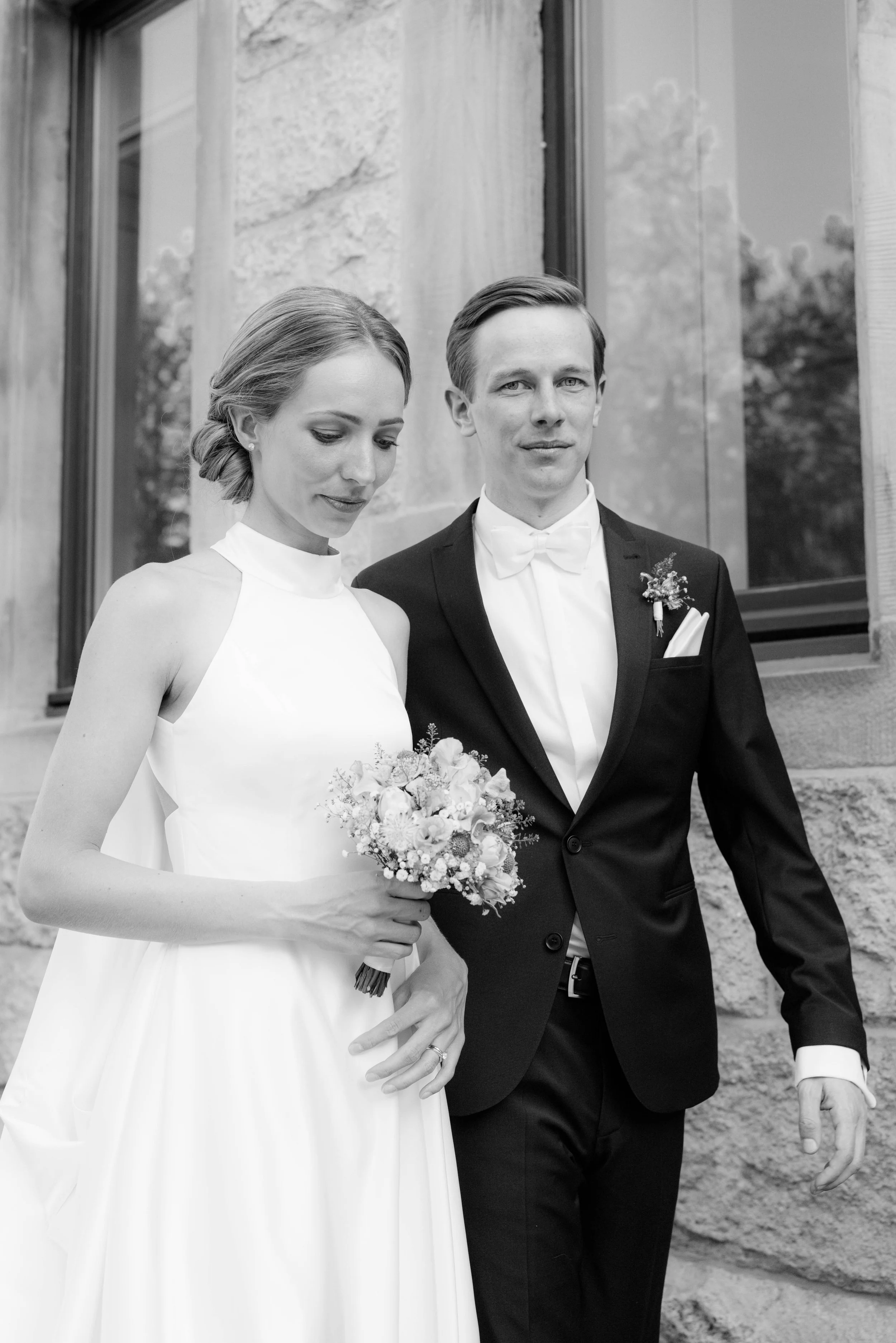 Schwarz-Weiß-Foto eines Hochzeitspaares, die vor einem Steingebäude stehen, die Braut hält einen Blumenstrauß und trägt ein weißes Brautkleid, der Bräutigam trägt ein schwarzes Sakko mit Fliege.