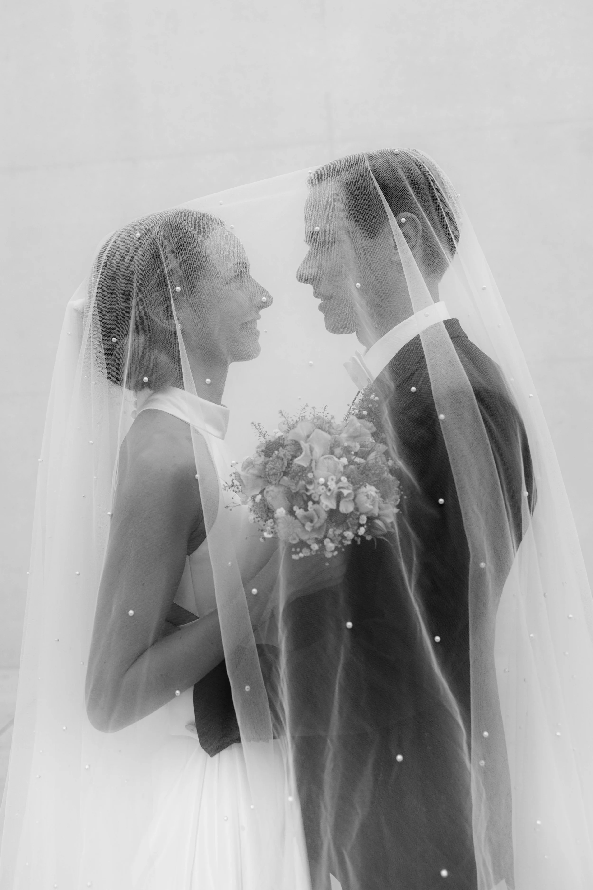 Ein Brautpaar steht sich unter einem durchsichtigen Schleier mit Perlen, die sich anlächelnd und mit dem Brautstrauß in der Hand gegenüberstehen.