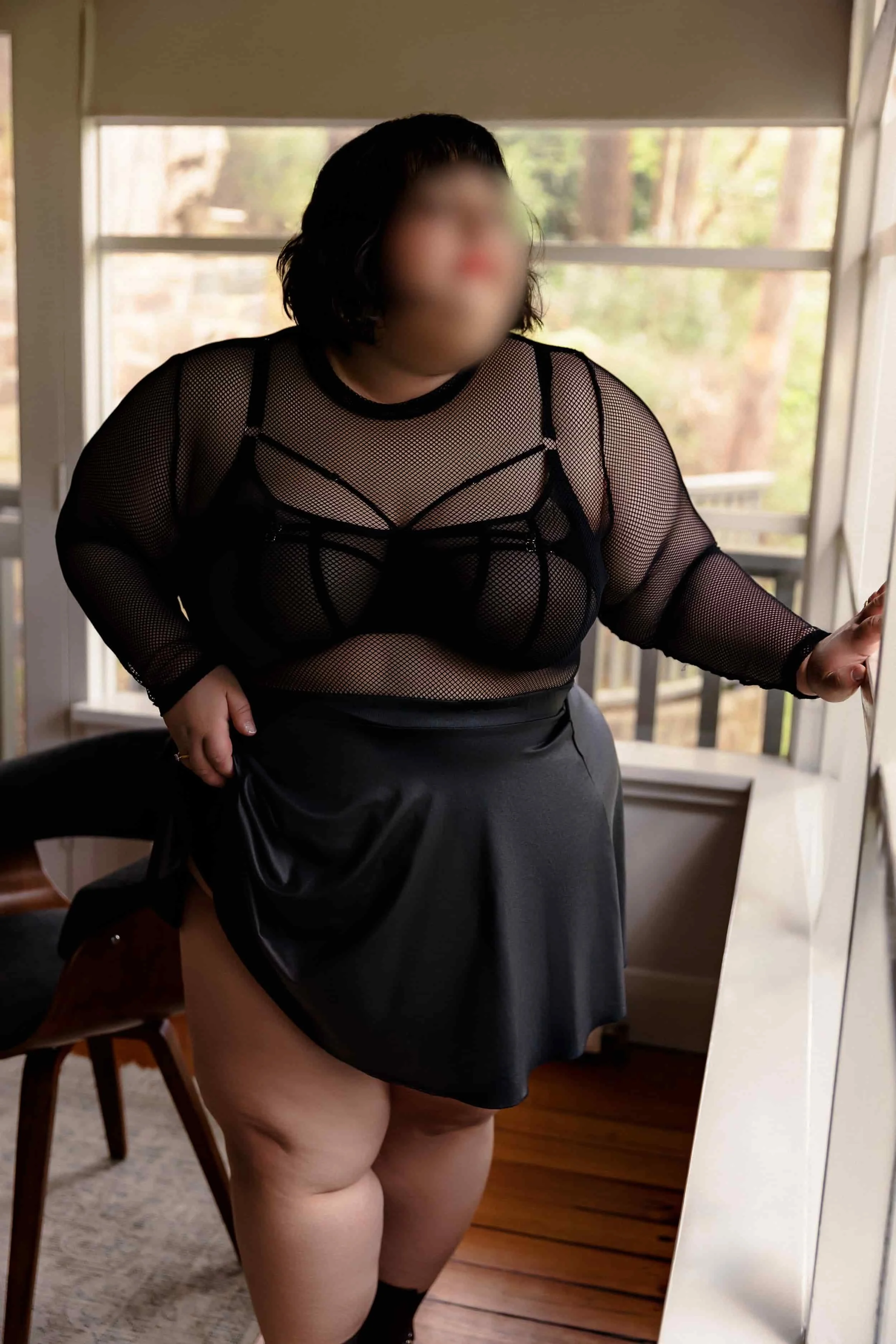 A woman with blurred face wearing a black mesh top and a pleather skirt, standing indoors near windows with outdoor trees visible in the background.
