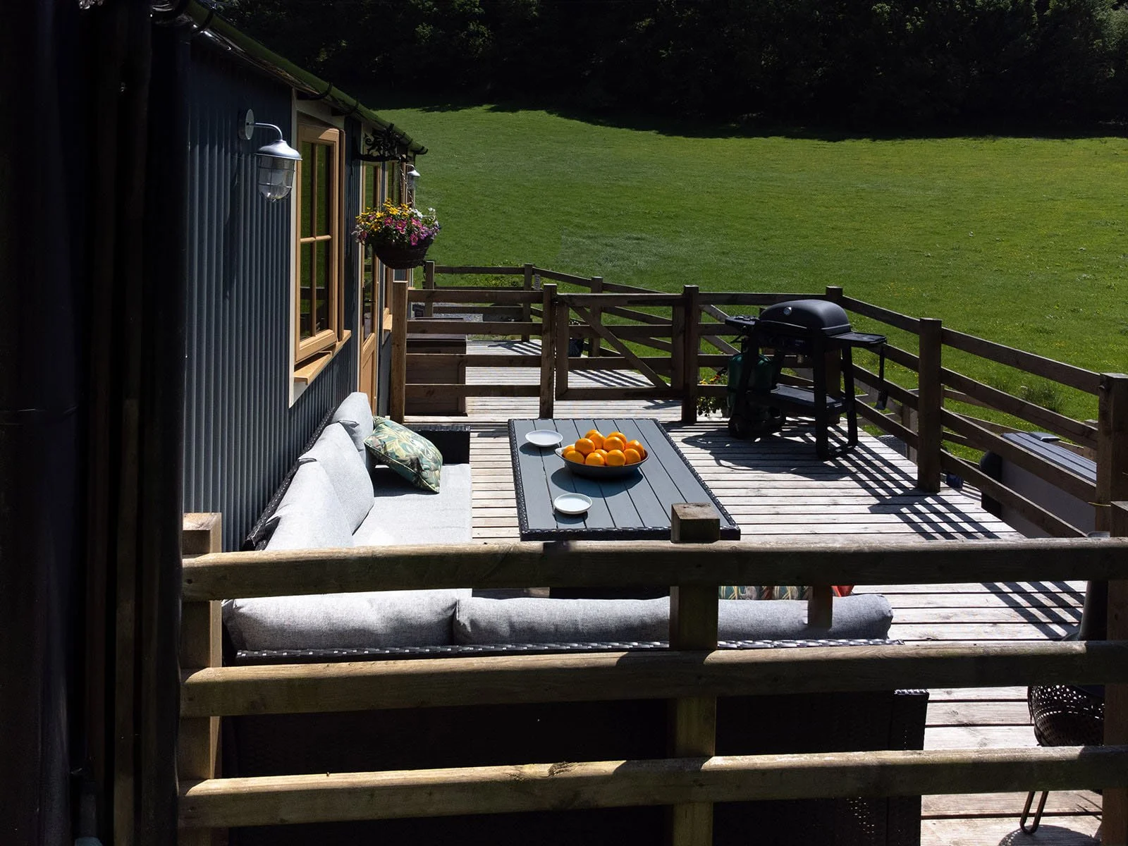 Backyard wooden deck with outdoor sofa, table with bowls of oranges, a barbecue grill, and lush green lawn in the background.