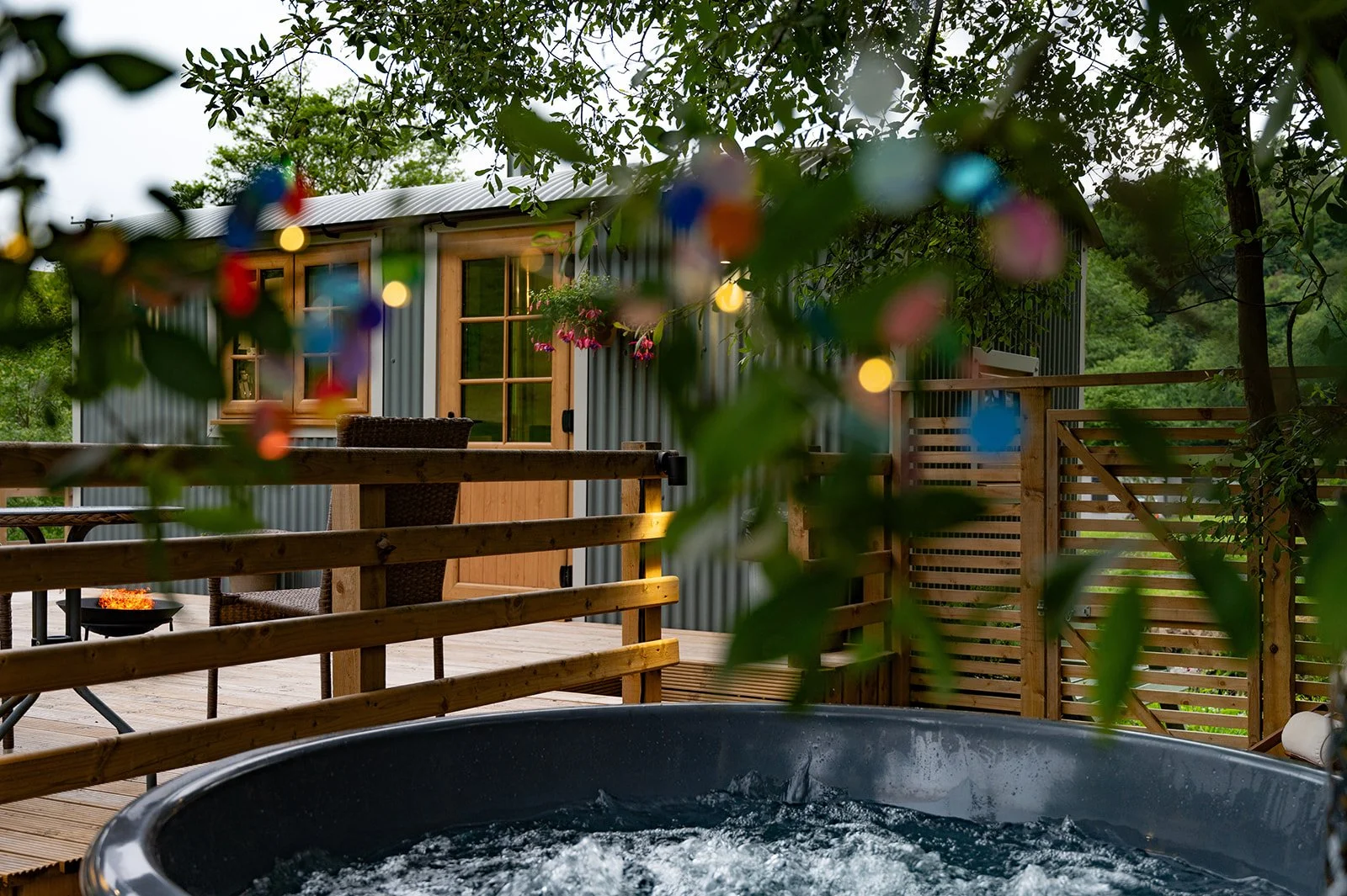 Outdoor wooden deck with hot tub, illuminated string lights, and a small building with a metal roof and wooden trim, surrounded by greenery and trees.