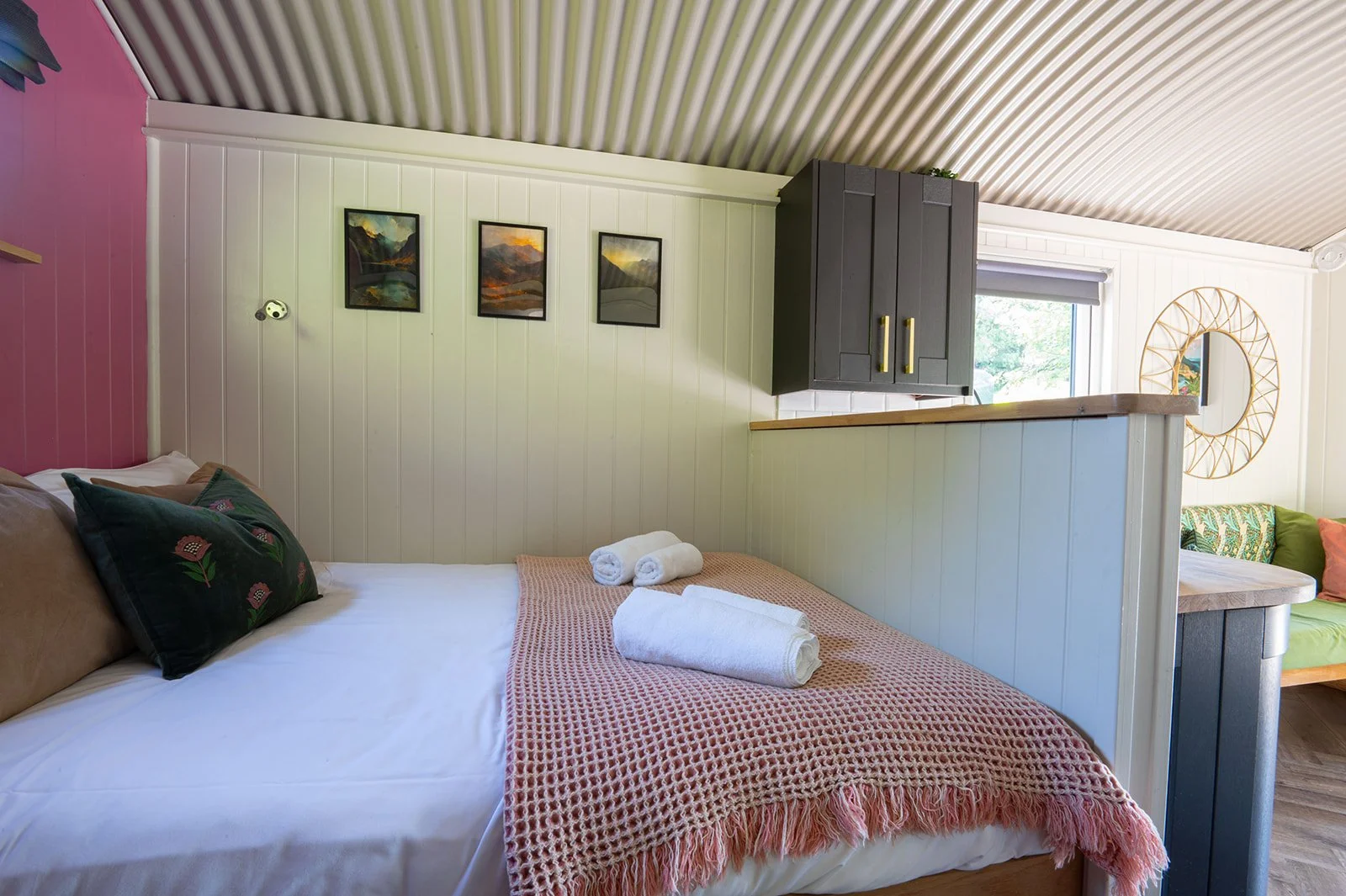 Queen-sized bed with a coral-colored crochet blanket, pillows, and rolled towels, in a cozy room with wall art and a sitting area with a round mirror.