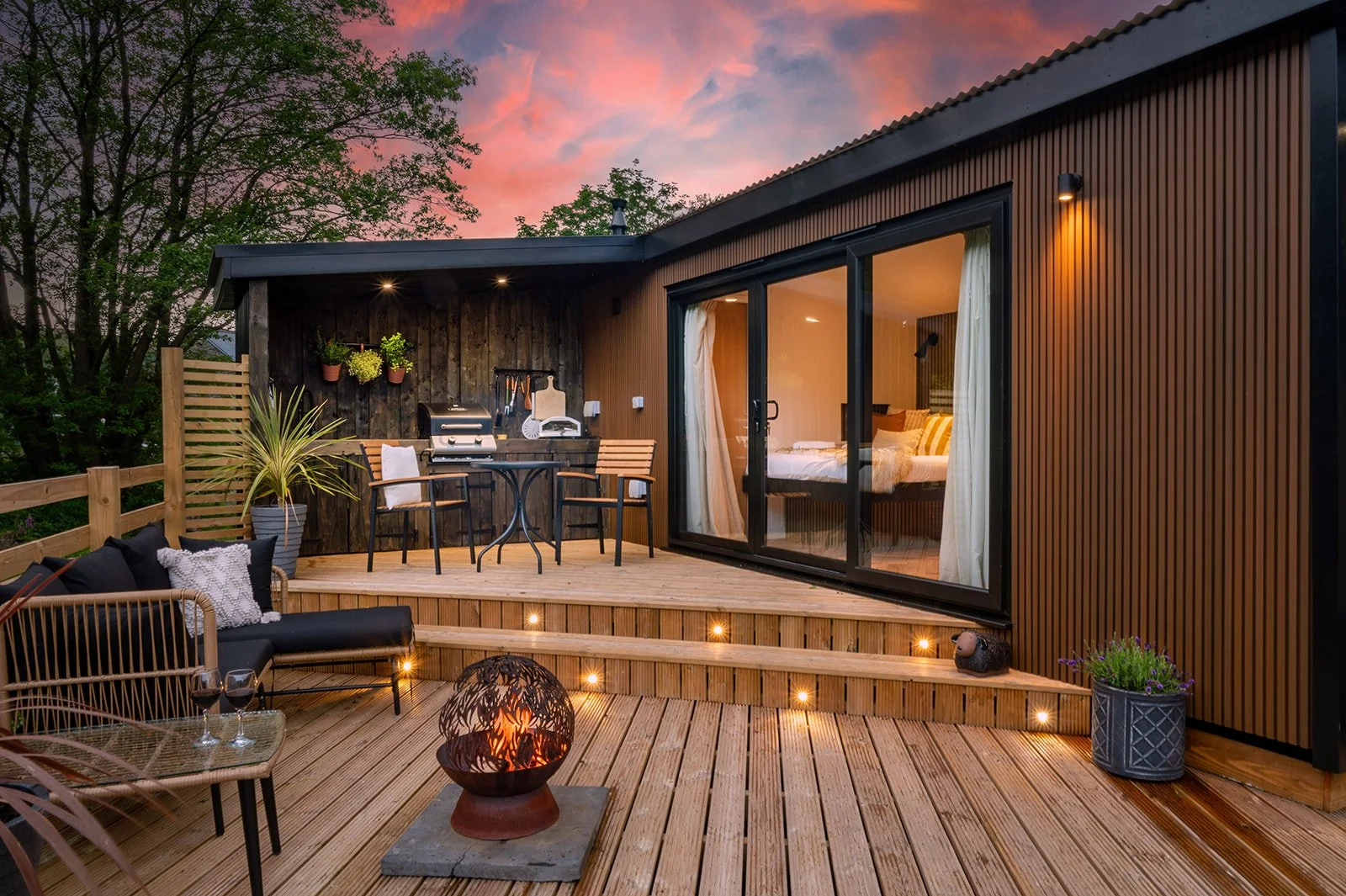 A cozy outdoor wooden deck with a built-in fire pit, patio furniture, potted plants, and a grilling station, overlooking a residential building with large glass doors leading to a bedroom, under a colorful sunset sky.