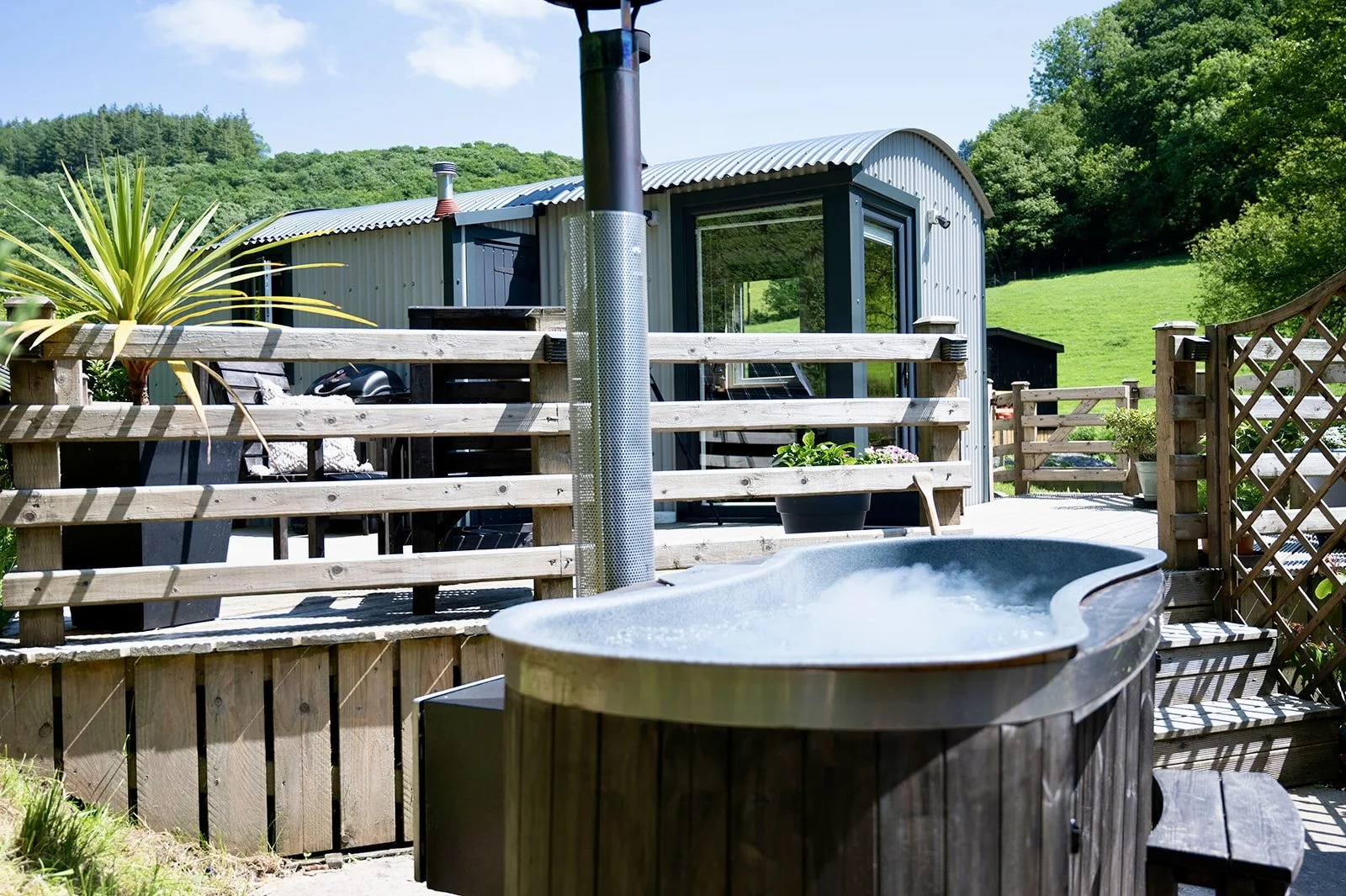 Outdoor deck with hot tub, wooden fencing, small garden with plants, metal shed, and lush green hills in the background on a sunny day.