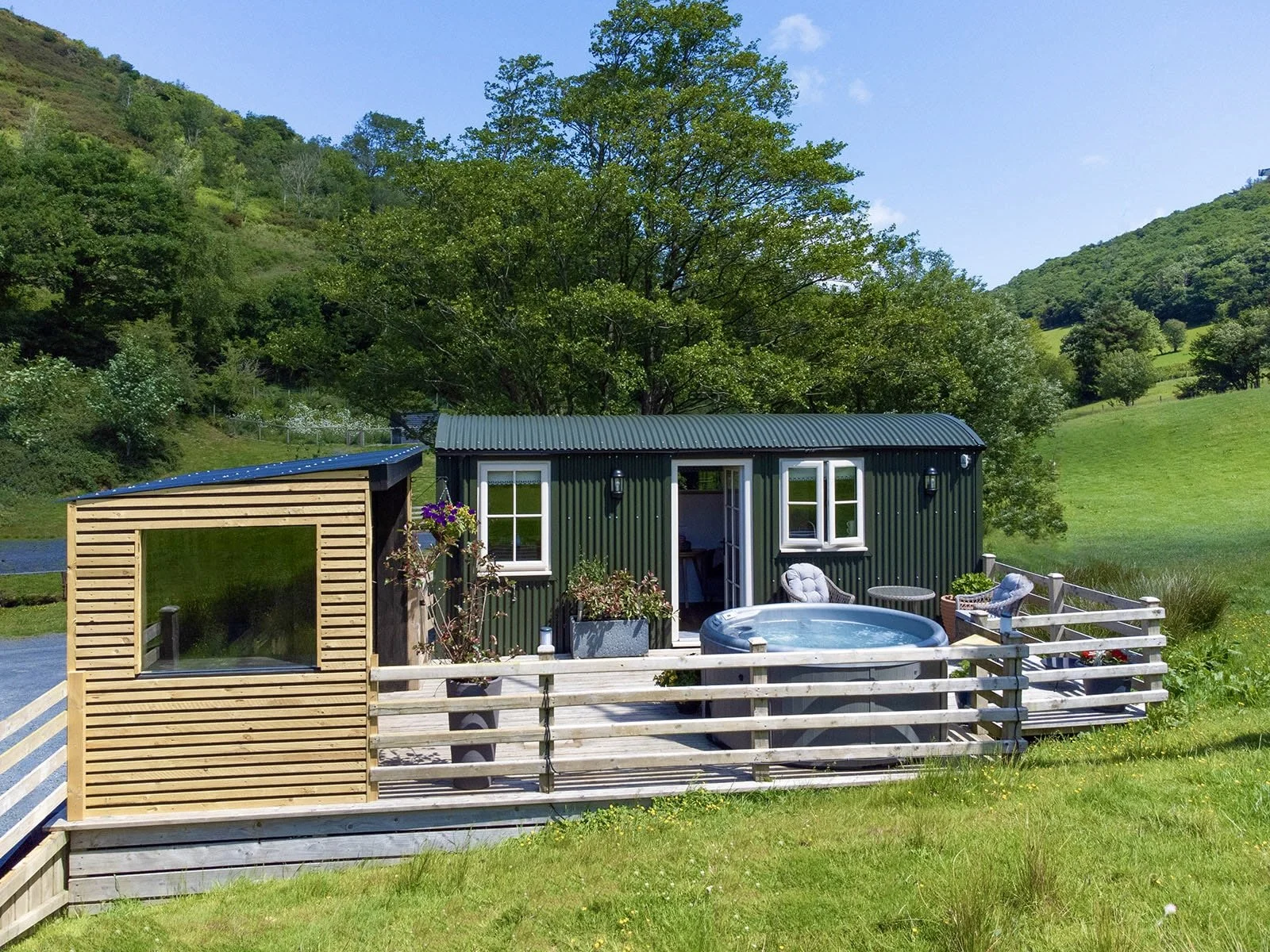 A green metal shepherds hut with white-framed windows in a lush, hilly landscape, featuring an outdoor deck with a hot tub, seating, and potted plants.