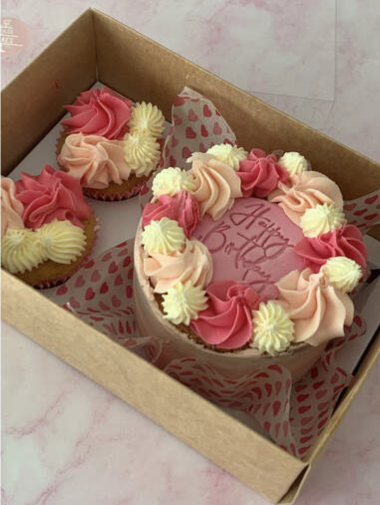 A birthday cake with pink and cream-colored frosting in a cardboard box, surrounded by four cupcakes with similar frosting, on a pink and white marble surface.