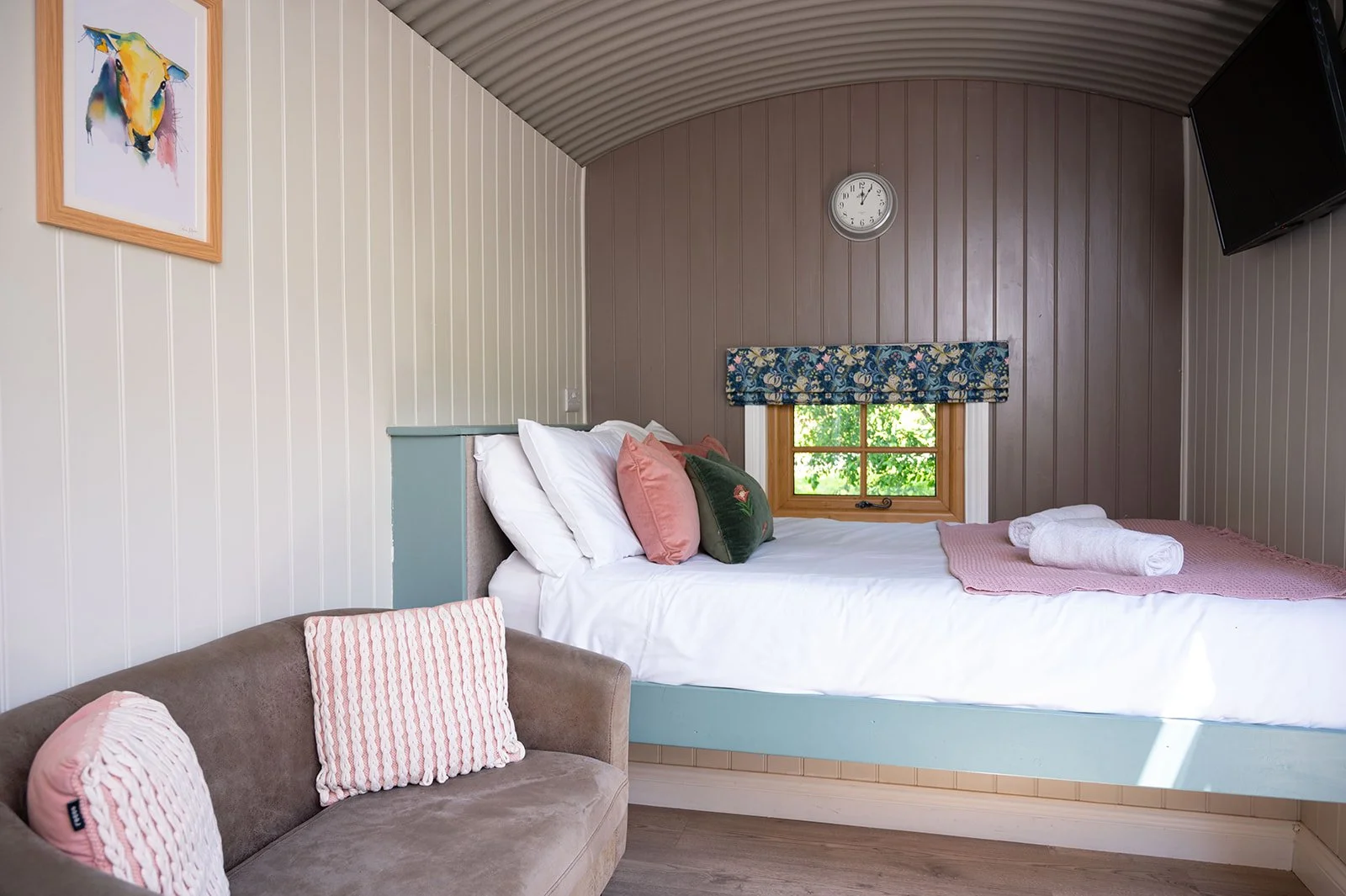 A cozy bedroom with a bed decorated with pink and green pillows, a small window with floral curtain, a hanging clock, a TV on the wall, a colorful cow portrait, and a beige sofa with pink and white pillows.