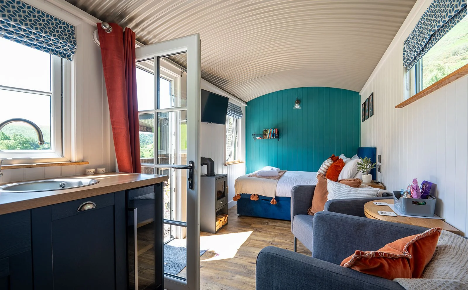 Cozy studio apartment with a bed, gray sofa, small table, and kitchen area with a sink and cabinet, decorated in blues, whites, and orange accents.