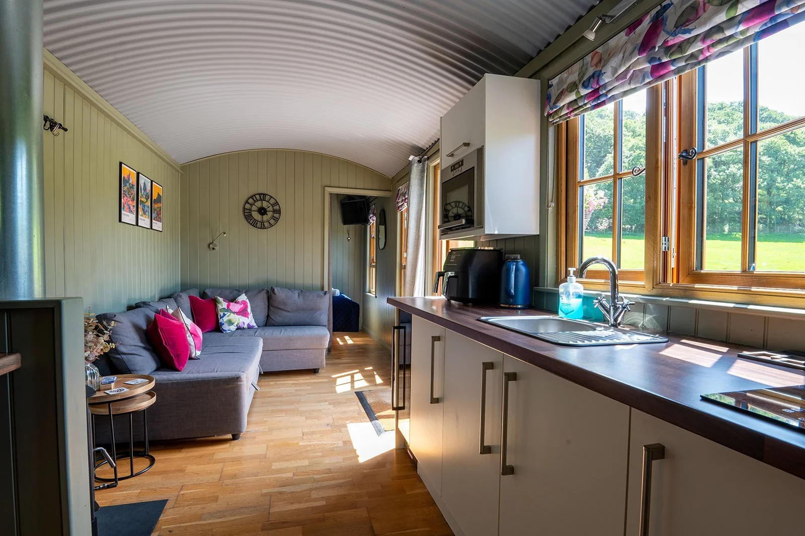 Interior of a cozy living space with a sofa, wall clock, and small side tables, adjacent to a kitchen area with a counter, sink, and large windows showing a green outdoor view.