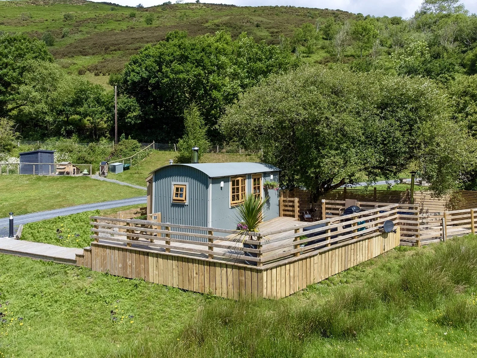A small, curved, metal shed with several windows, situated on a wooden deck, surrounded by a wooden fence, and adjacent to a large leafy tree in a rural area with green grass and rolling hills in the background.