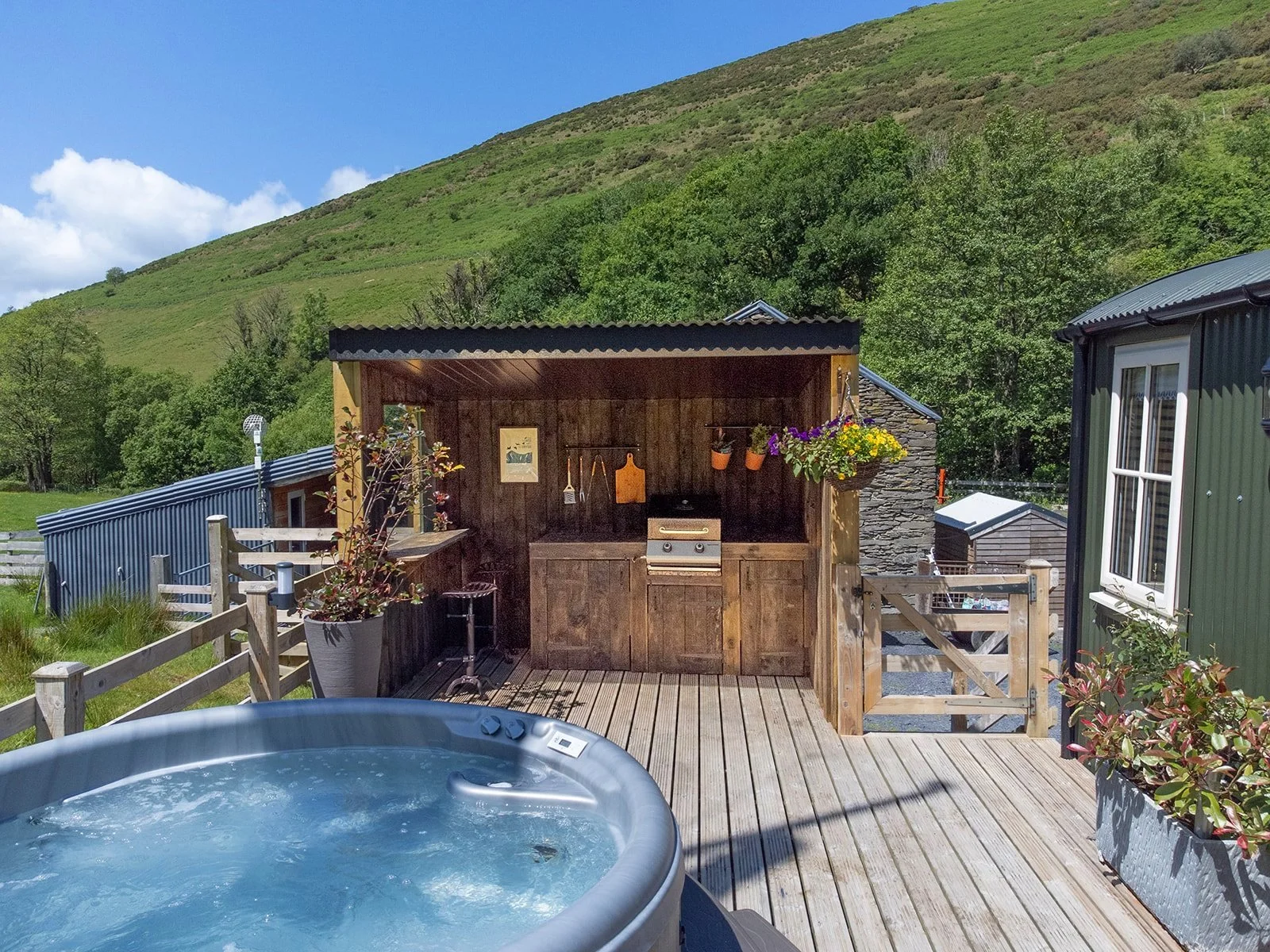 Outdoor deck with hot tub, wooden shed with barbecue, potted plants, hanging flower basket, in a green hillside landscape with blue sky.
