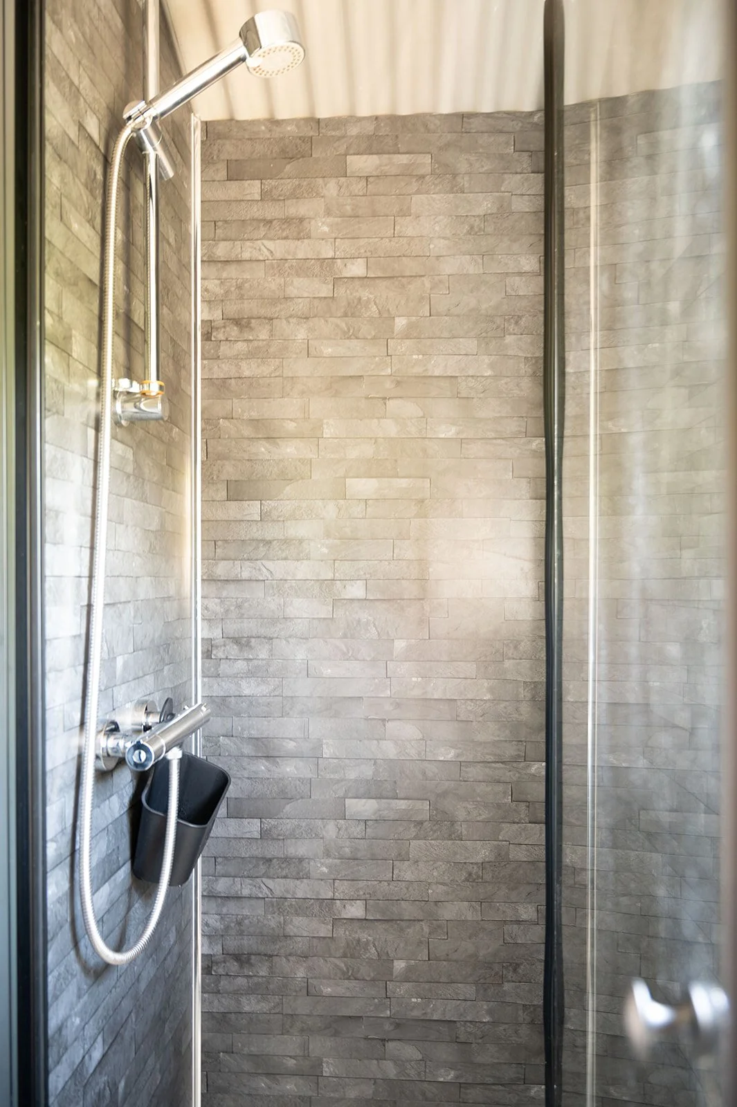 Empty modern shower with gray brick wall tiles, a silver showerhead, and a black soap holder.