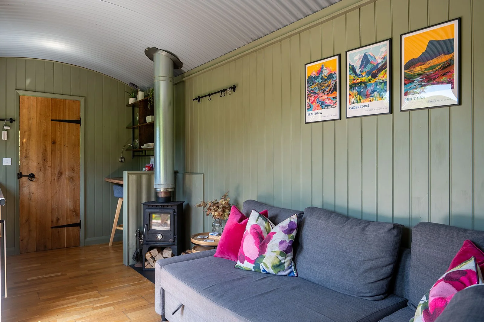 Living room with green paneled walls, wooden floor, a gray sofa with pink and floral pillows, a small wood stove, shelves with books and decor, and framed colorful mountain landscape artwork.