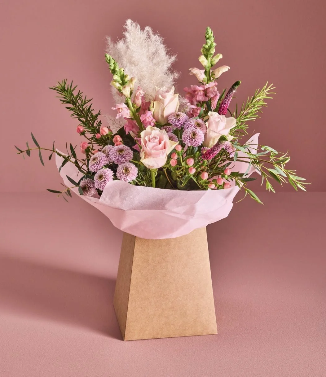 A bouquet of pink flowers including roses, chrysanthemums, and snapdragons, with green foliage, wrapped in pink tissue paper in a plain brown paper bag, set against a pink background.