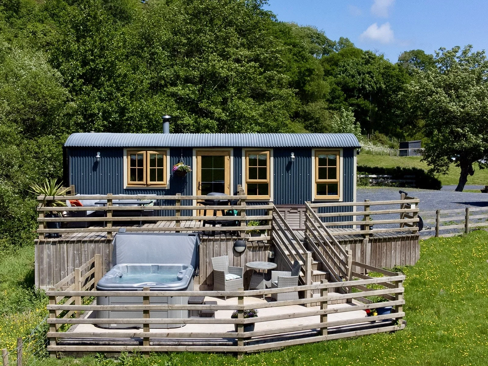 Blue tiny house with wooden trim, multiple windows, and a deck area with outdoor furniture and hot tub, surrounded by a wooden fence, green grass, trees, and a gravel driveway, under a clear blue sky.