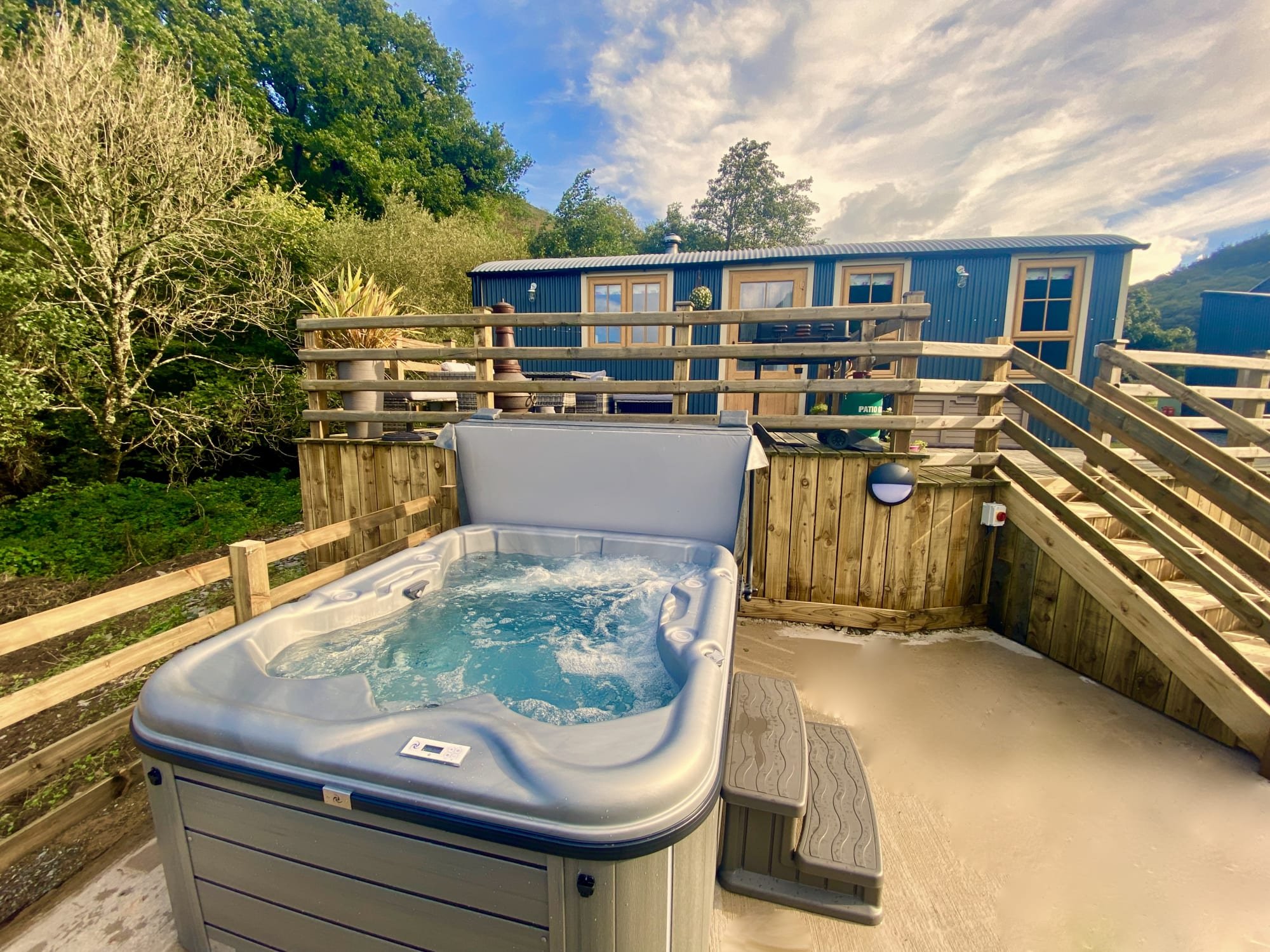 Outdoor wooden deck with a hot tub filled with bubbling water and a small set of stairs, surrounded by trees and a blue building in the background under a partly cloudy sky.