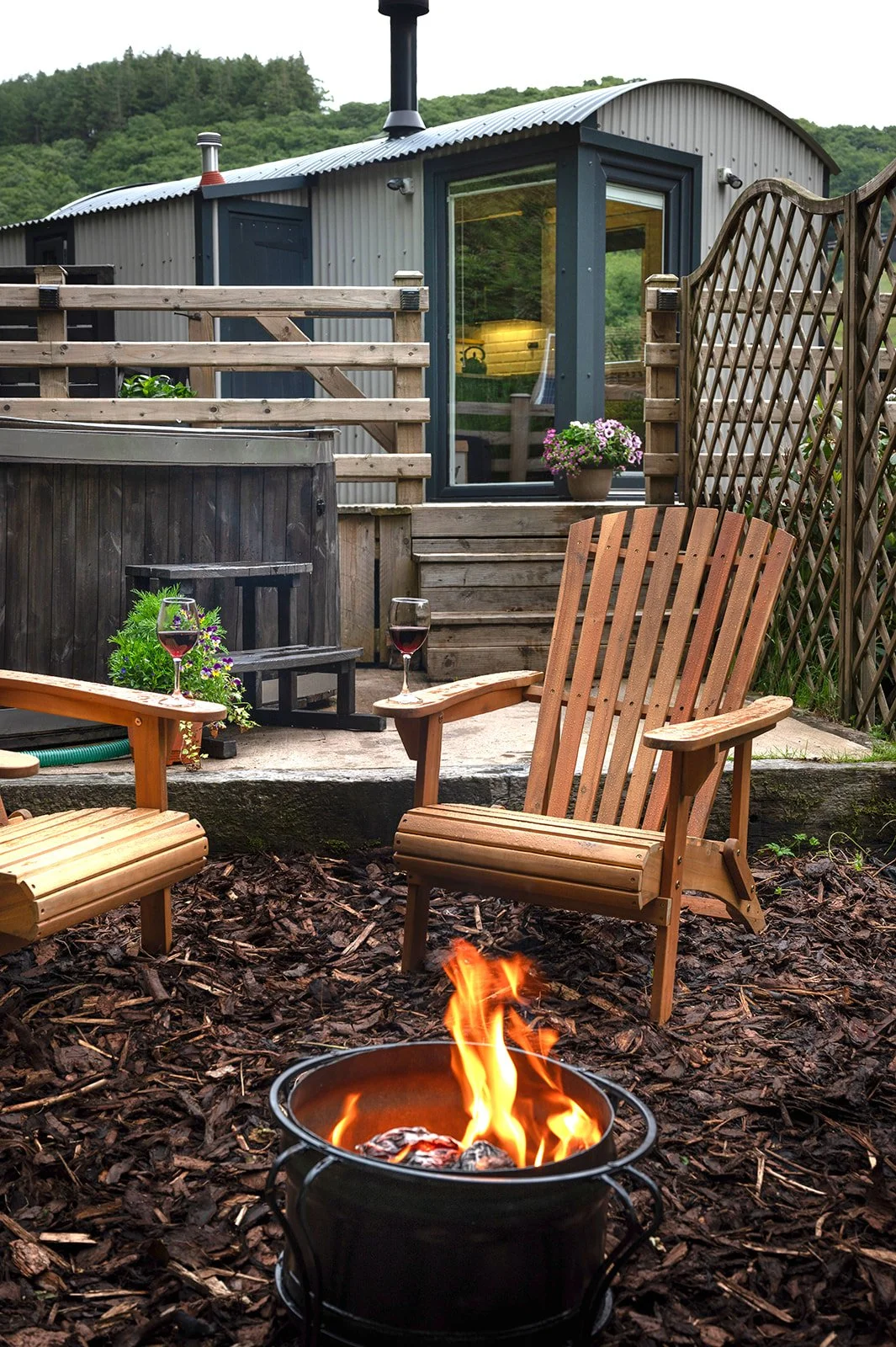 A cozy outdoor patio with wooden chairs, a fire pit, and wine glasses, overlooking a small building with a metal exterior, surrounded by lush green hills.