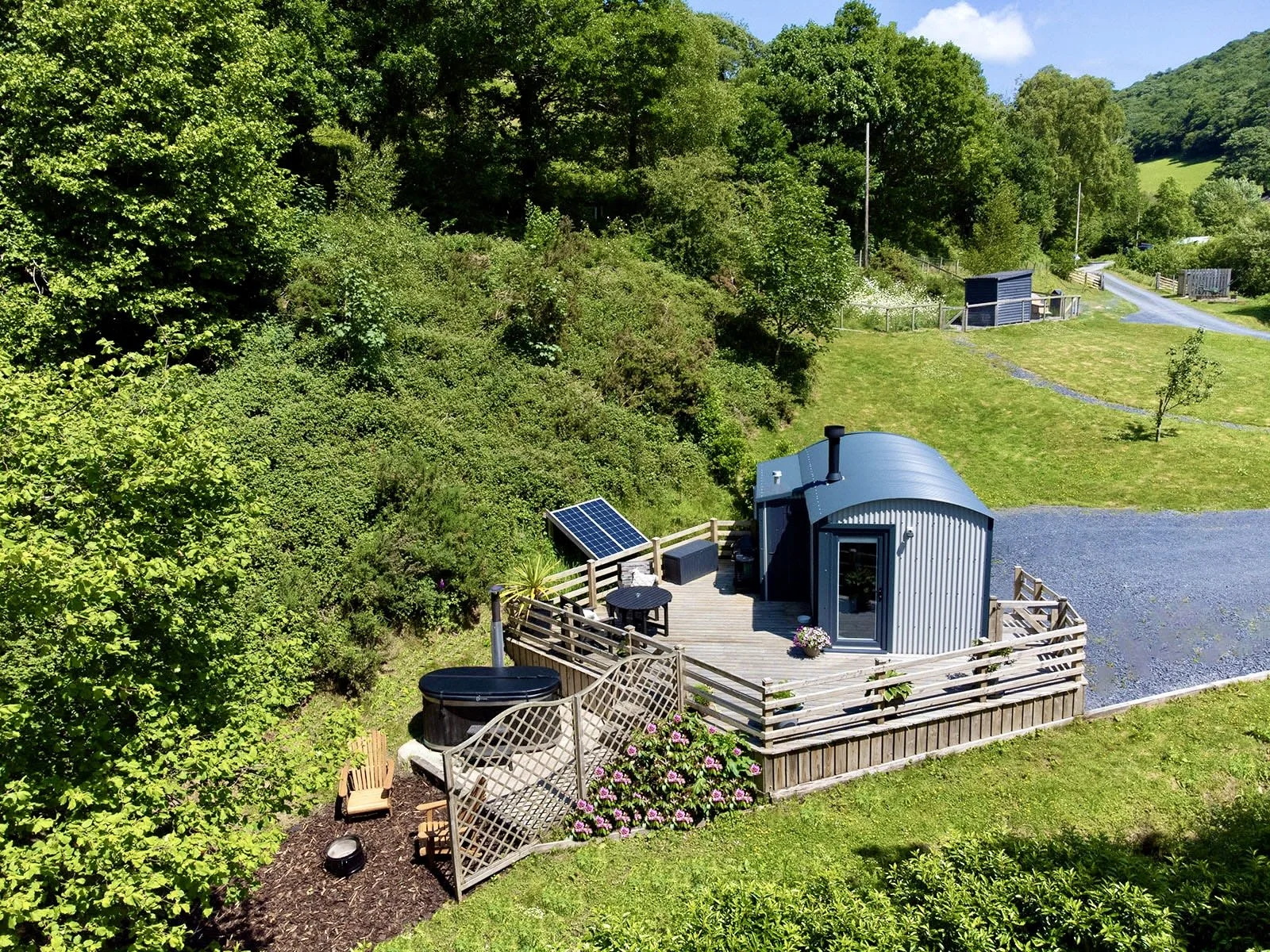 A tiny blue cabin with a curved roof on a wooden deck surrounded by green trees and plants, with a hot tub, solar panel, and outdoor furniture.
