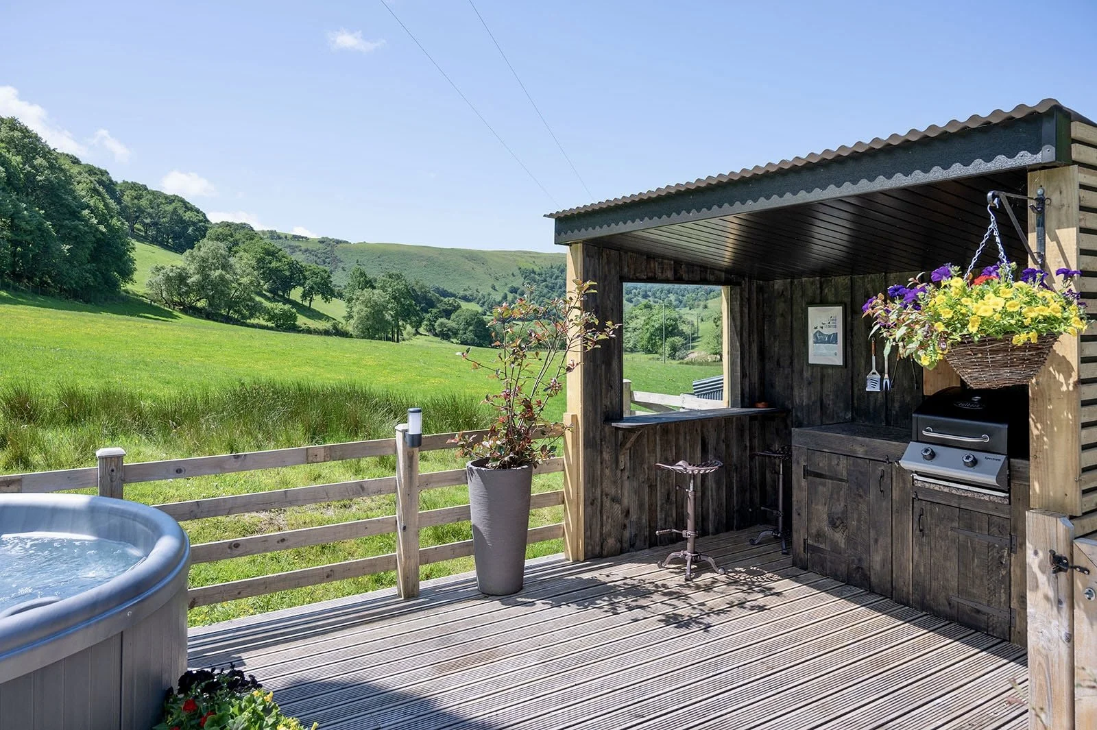 Outdoor deck with hot tub, potted plants, a small outdoor kitchen with a grill, hanging flower basket, and scenic green countryside with hills and trees under a blue sky.