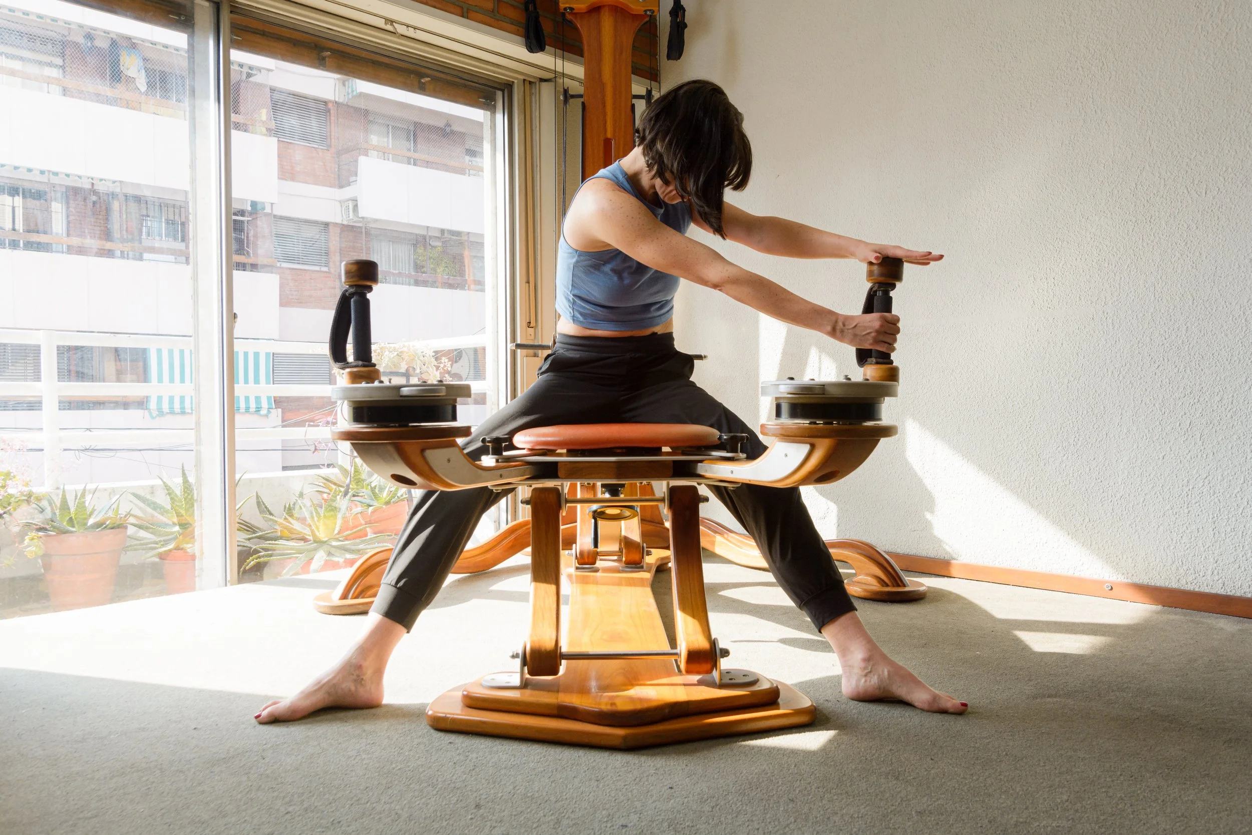 Woman exercising on a wooden flexibility machine by a large window.