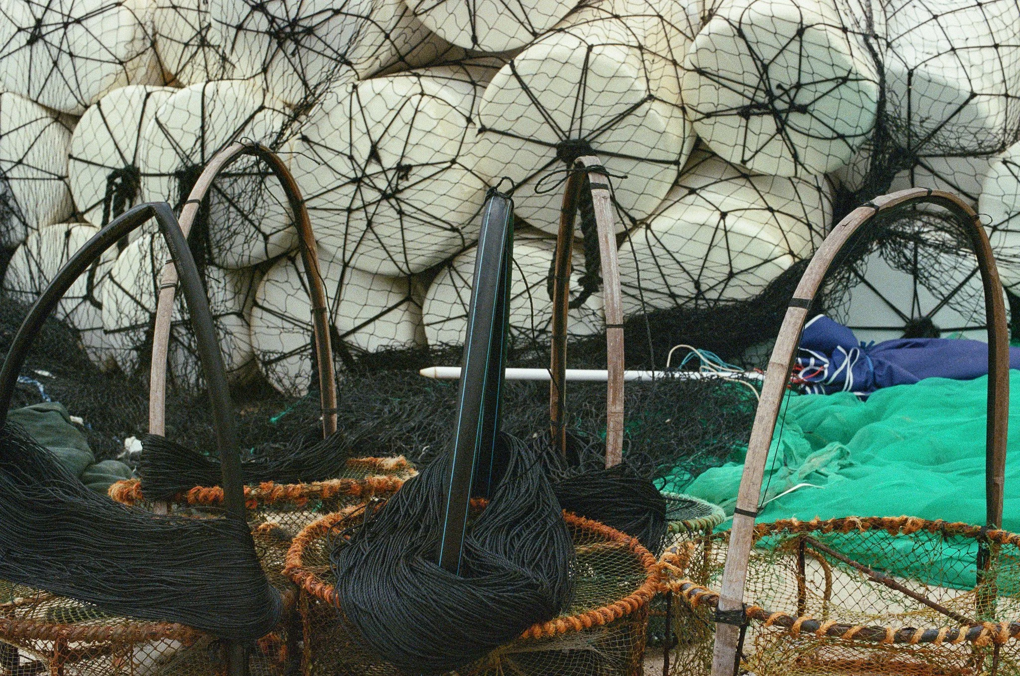 Various fishing nets, floats, and equipment stacked together, including black and orange nets, some upright and some inside baskets, with large white floats secured in a black net in the background.