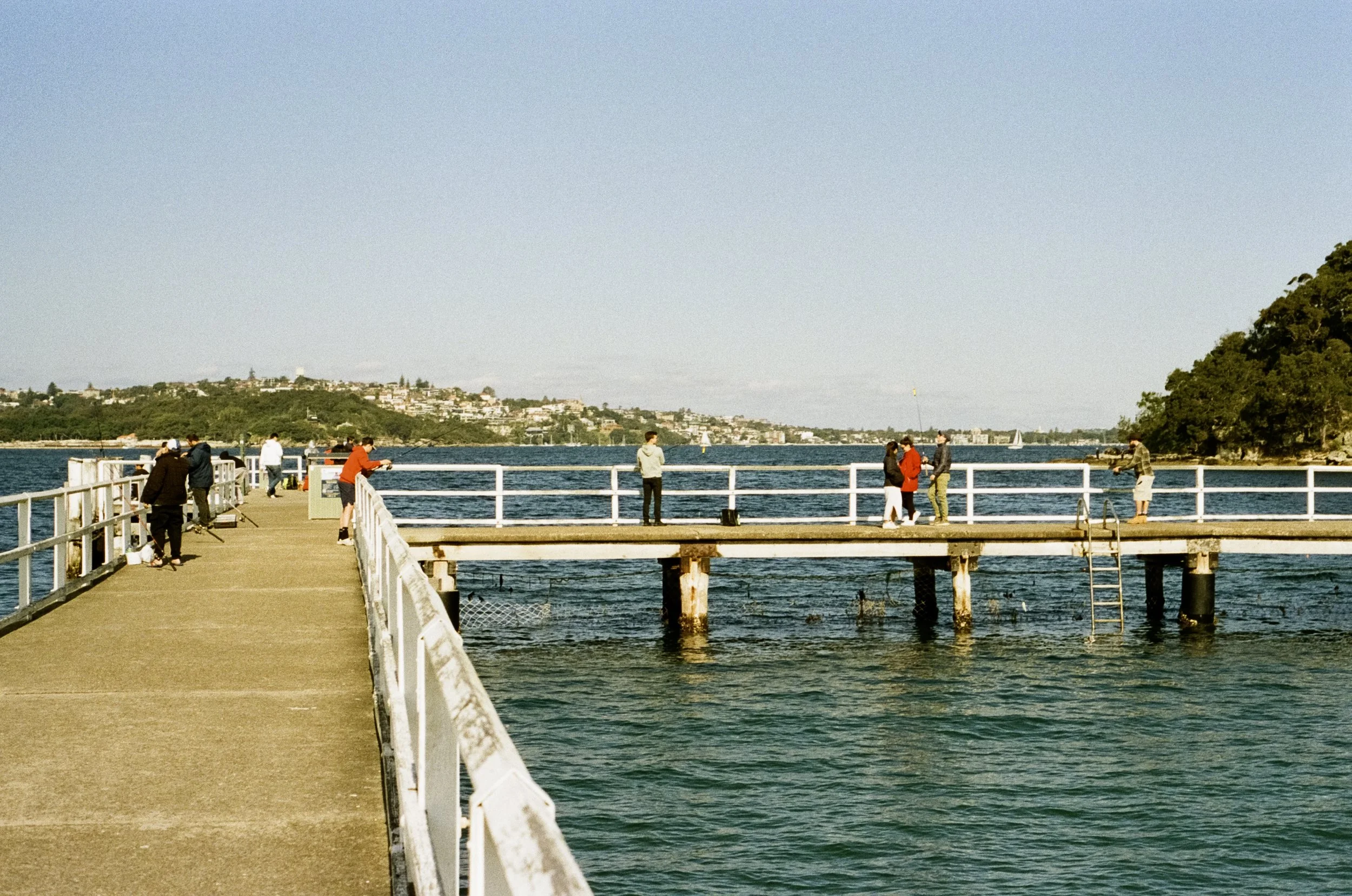 People fishing and walking on a wooden pier extending into a body of water, with a distant shoreline and trees on the right.