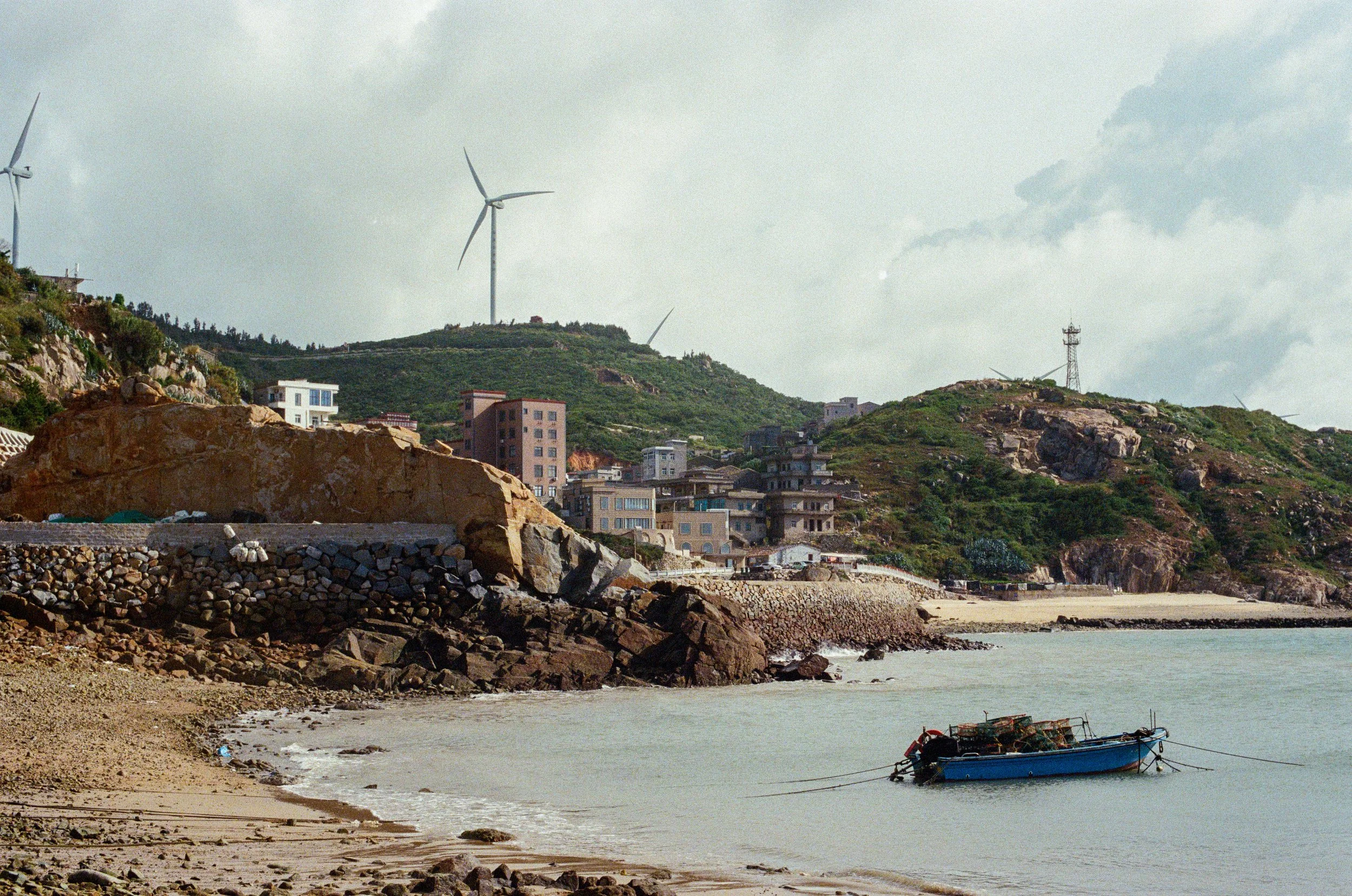 A coastal scene with a small boat floating on calm water, sandy beach in the foreground, rocky shoreline, uphill green hills with scattered buildings, and wind turbines on the hilltops.