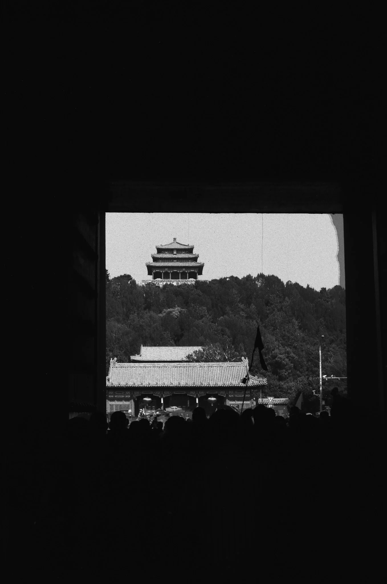 Silhouettes of people looking out an open window toward a traditional Asian pagoda on a hilltop, with trees and a building with a tiled roof in the foreground.