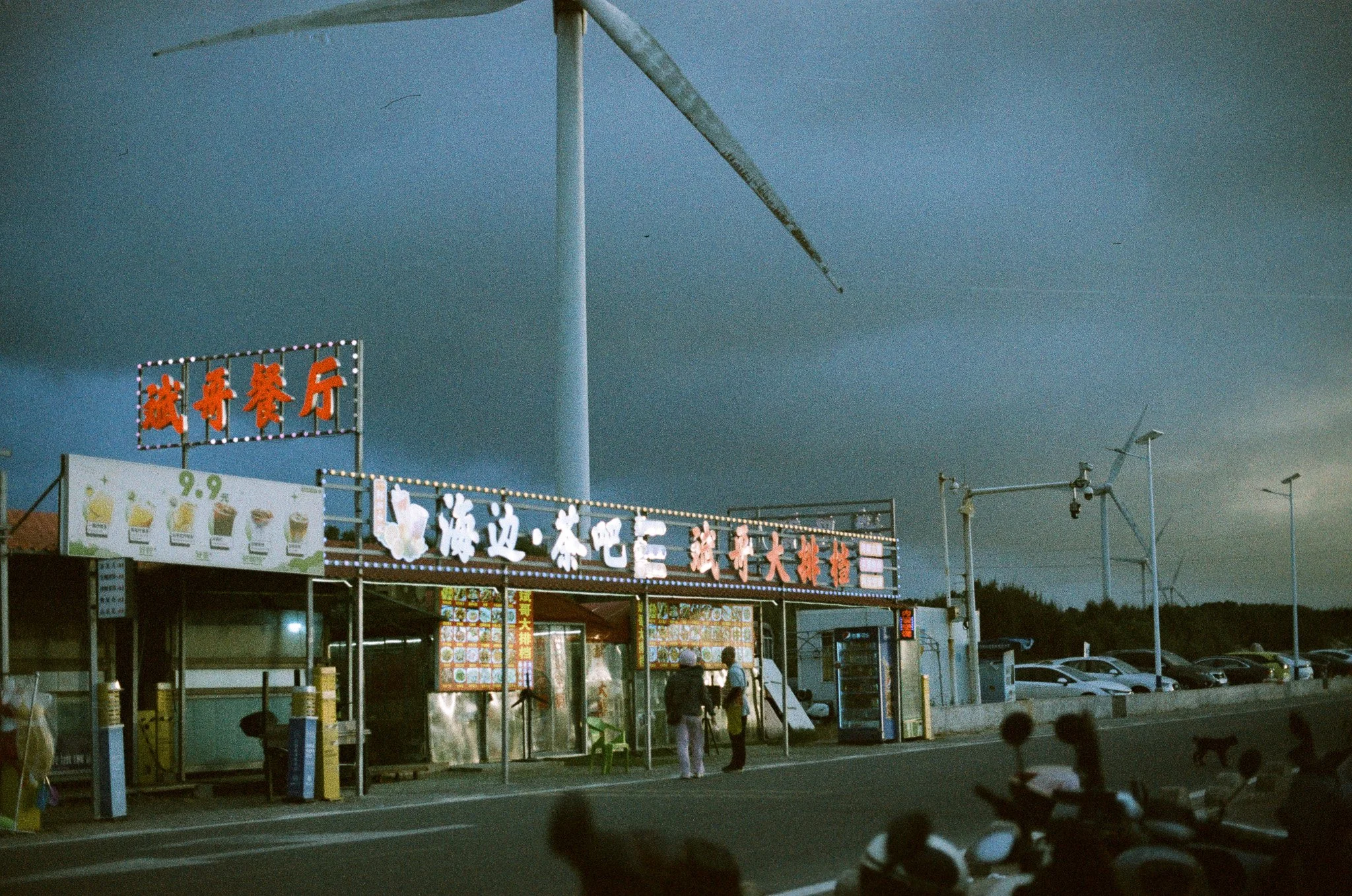 A street scene with a restaurant and a parking lot with several cars. The restaurant has bright neon signs with Chinese characters and images of drinks. Two people are standing and talking in front of the restaurant, with dark, stormy clouds overhead.