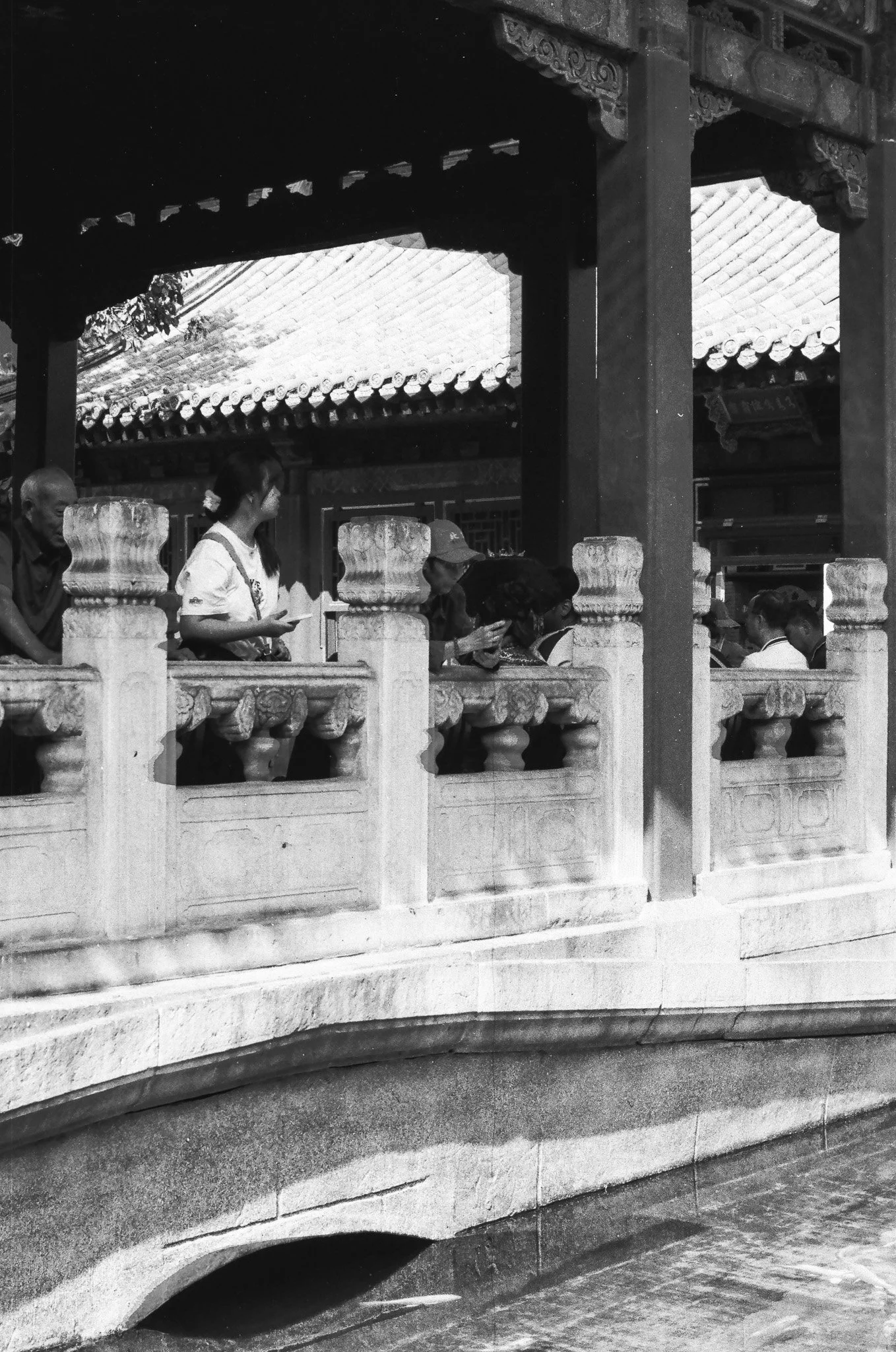 People standing on a decorative stone bridge with traditional Chinese architecture, including tiled roofs and intricate woodwork.