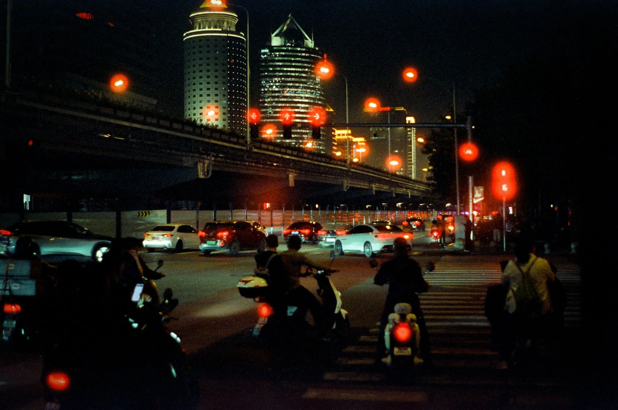 Nighttime city street scene with several vehicles and people on scooters at a crosswalk, illuminated red traffic lights, and tall buildings in the background.