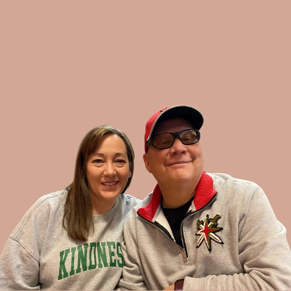 A woman and a man smiling and sitting close together against a plain background. The woman has shoulder-length brown hair and wears a gray sweatshirt with green lettering that says 'KINDNESS'. The man wears glasses, a red cap, and a gray zip-up jacket with a patch featuring a star and a sword.