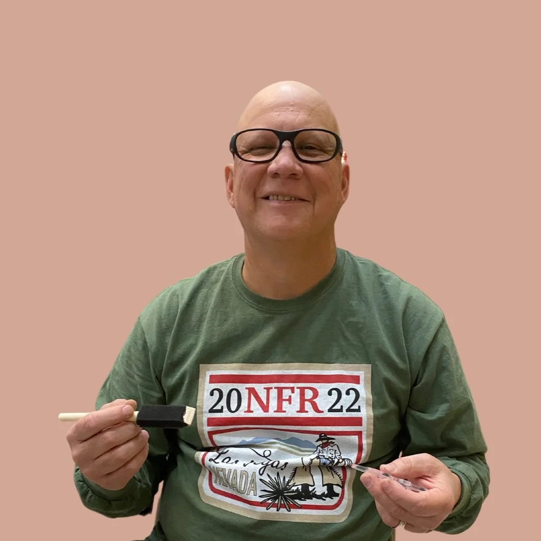 A smiling bald man wearing glasses and a green shirt with Nevada-themed print, holding a sushi roller in one hand and a small plastic packet in the other, against a plain pink background.