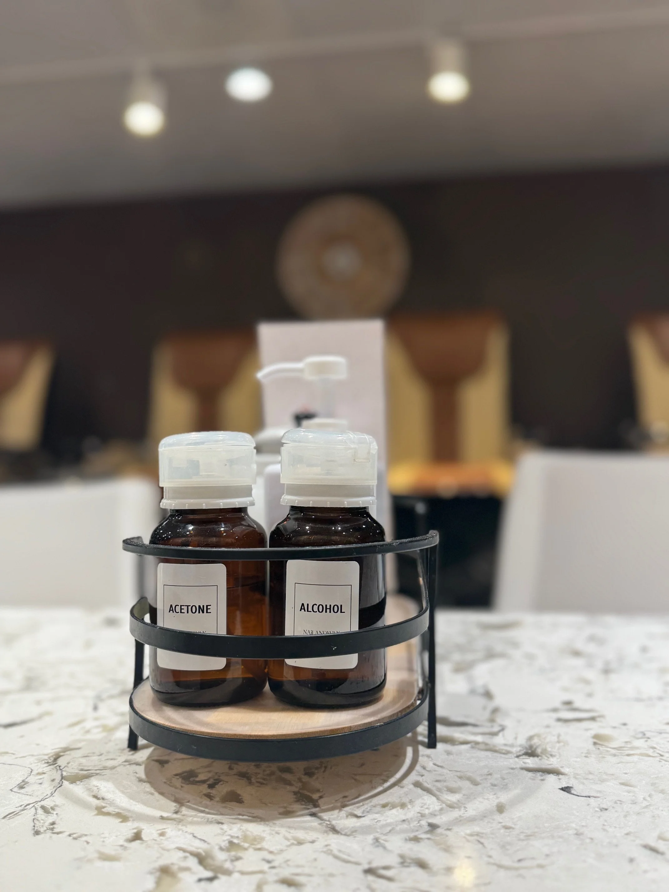 Two bottles labeled "Acetone" and "Alcohol" on a marble countertop, placed in a circular black holder, with a blurred salon background.