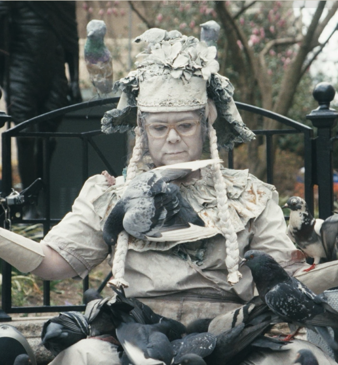 A person dressed in an elaborate costume with feathers and fabric, wearing glasses, and surrounded by pigeons, sitting on a park bench with trees in the background.