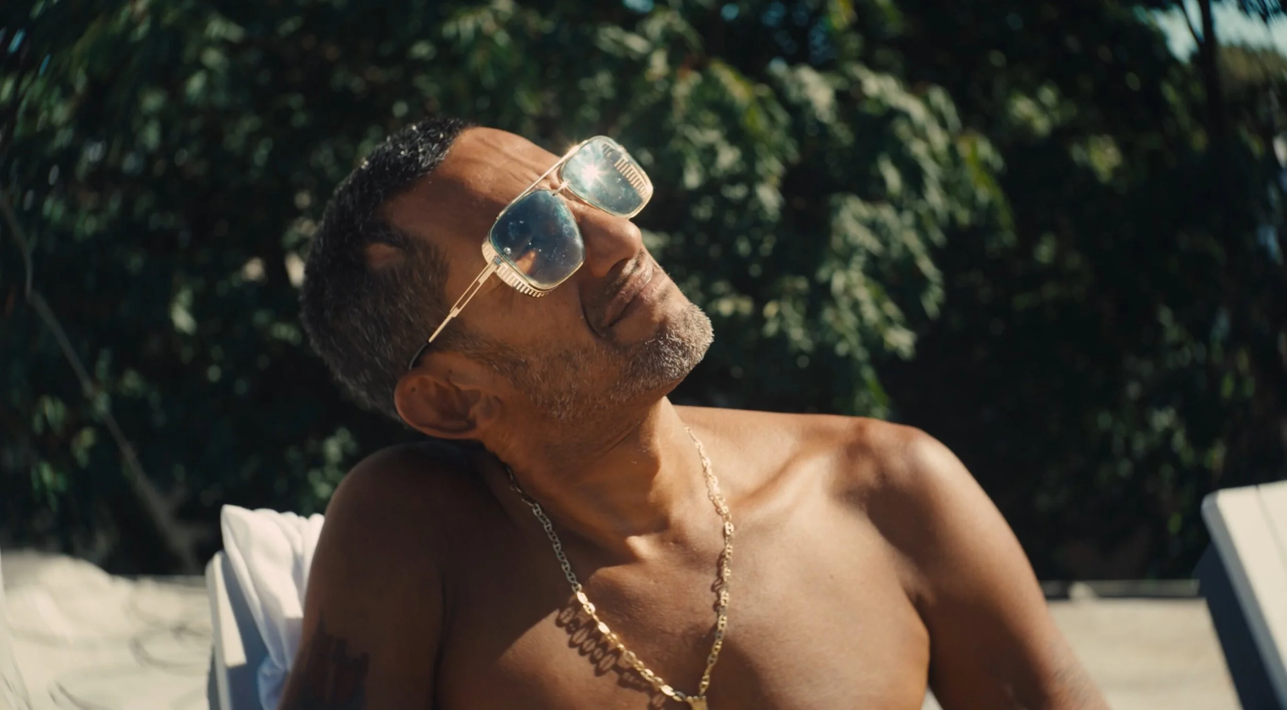 A shirtless middle-aged man relaxing outdoors in sunlight, wearing sunglasses and a gold chain, with a background of green trees.