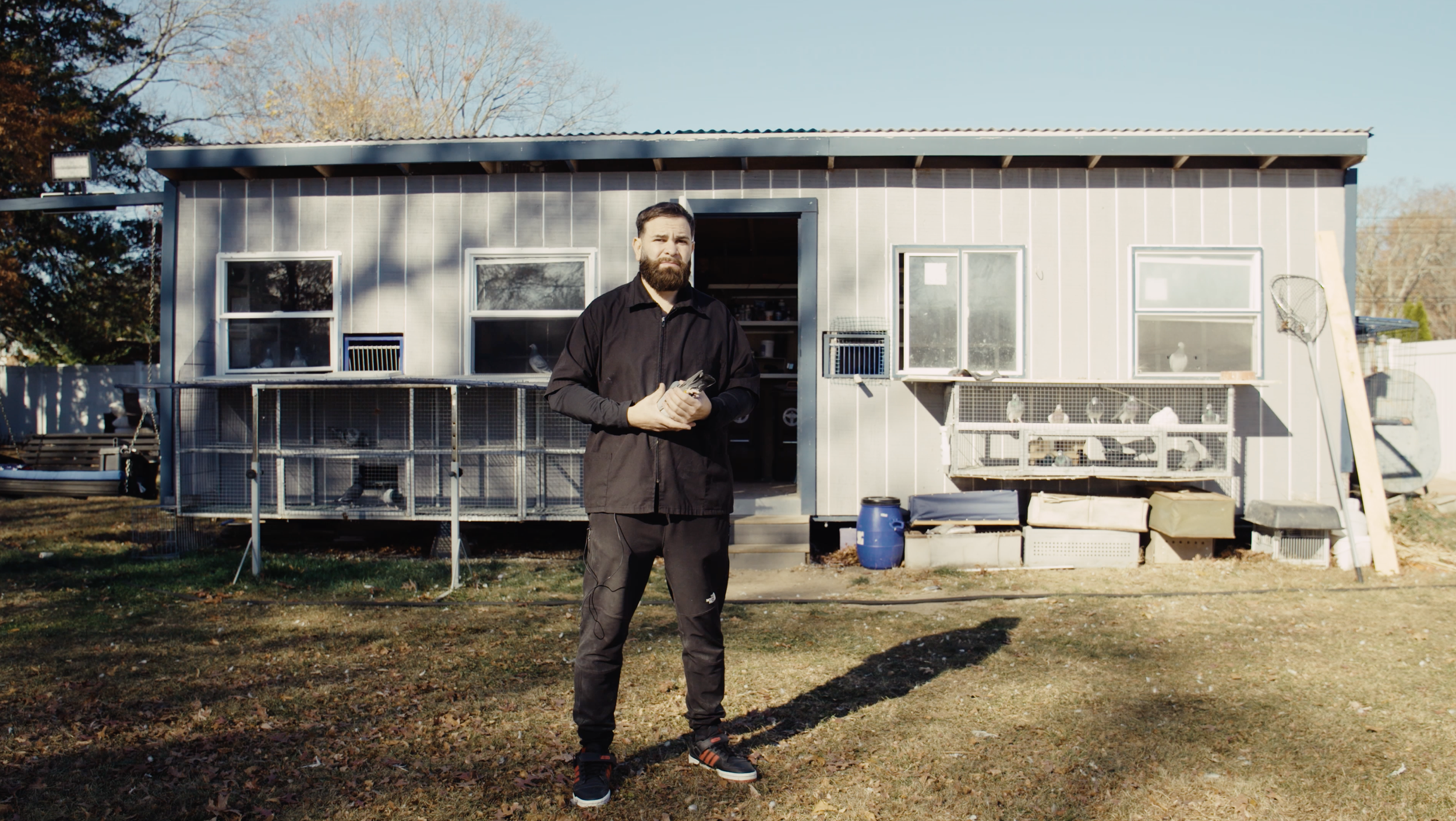 A man with a beard standing in front of a house with many windows, holding a pigeon, with outdoor cages and equipment visible.