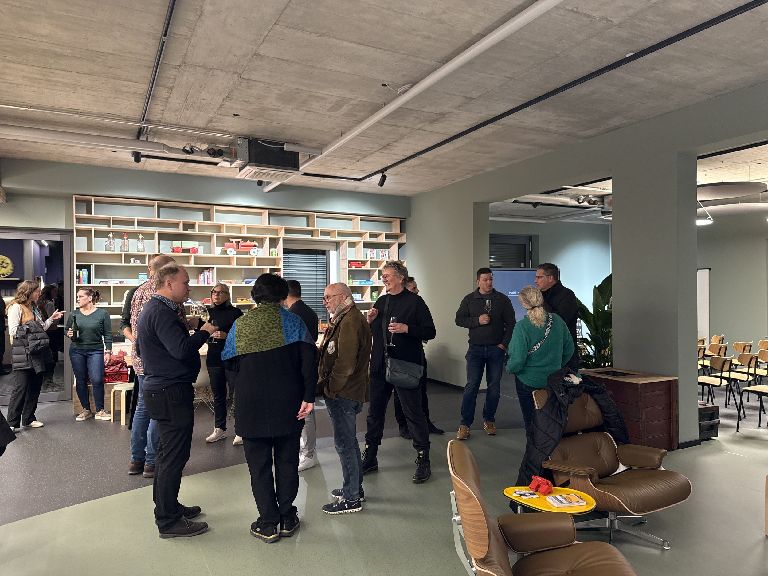 Menschen bei einer gesellschaftlichen Veranstaltung im Innenraum, einige trinken Wein, einige sprechen miteinander, im Hintergrund Bücherregal und Stühle.