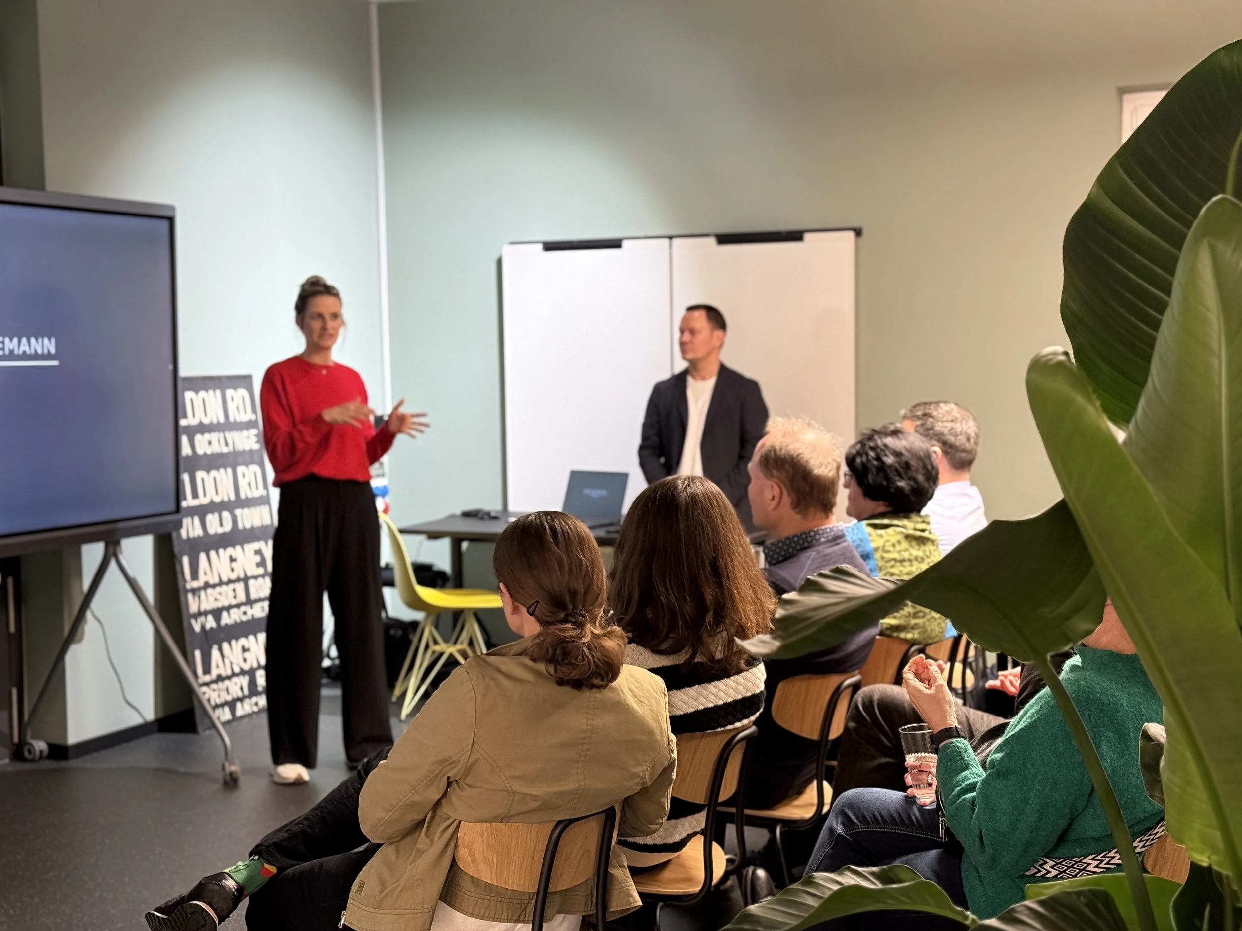 Eine Gruppe von Menschen hört einem Vortrag zu, während eine Frau in rotem Oberteil spricht und ein Mann im schwarzen Sakko daneben steht, im Raum mit weißen Wänden und einem großen Bildschirm.
