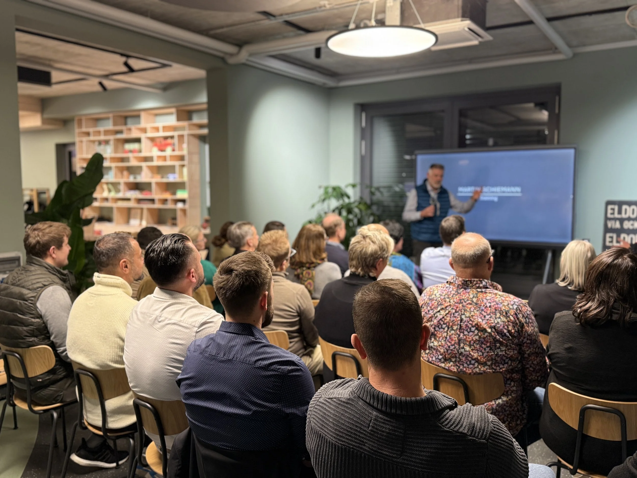 Menschen in einem Konferenzraum, die einer Präsentation auf einem großen Bildschirm zuhören. Der Referent steht vor dem Bildschirm und zeigt auf die Folie.