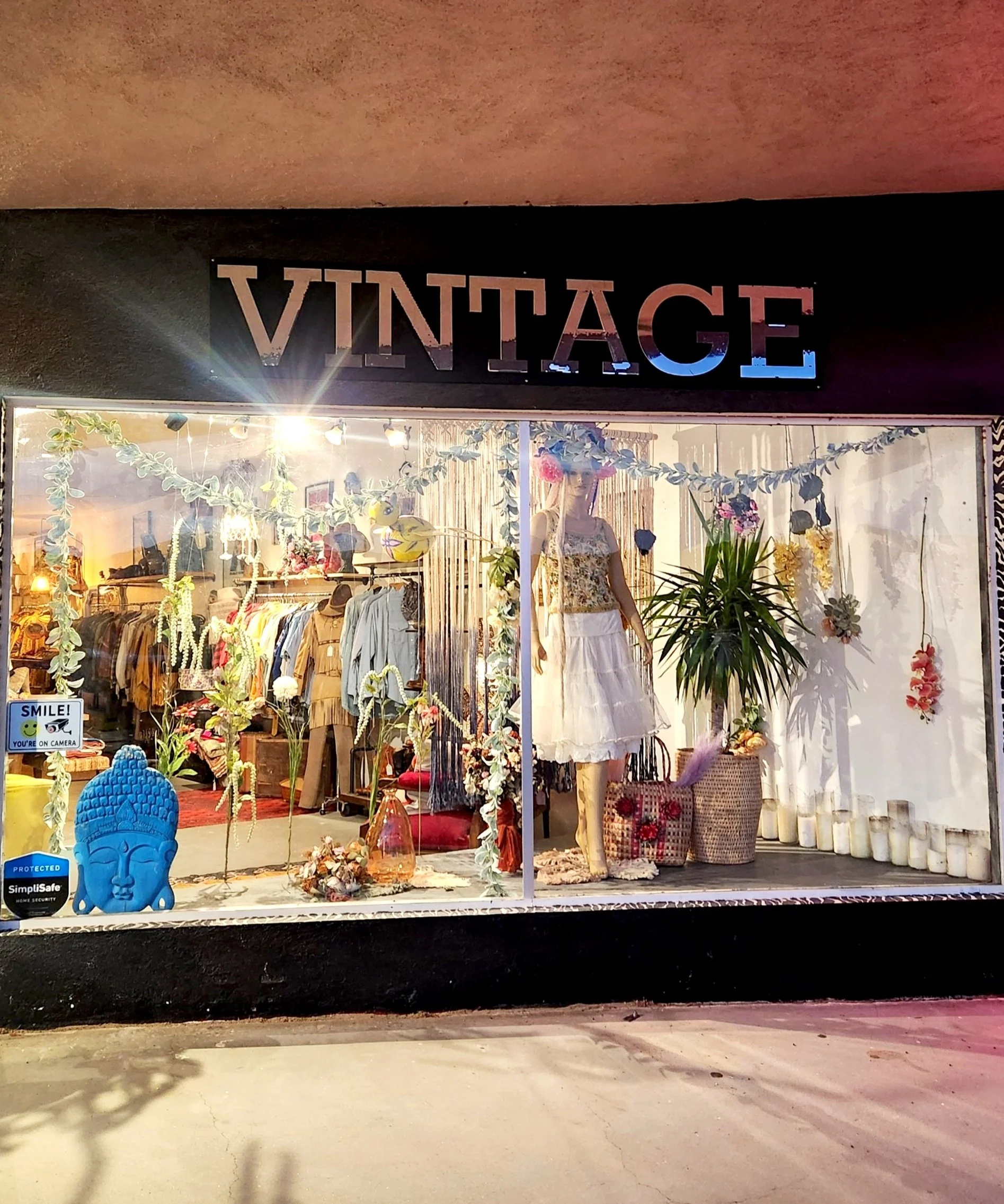Front of a vintage store with clothing displayed, a mannequin wearing a dress, decorative plants, a blue Buddha statue, and a "Smile! You're on Camera" sign.
