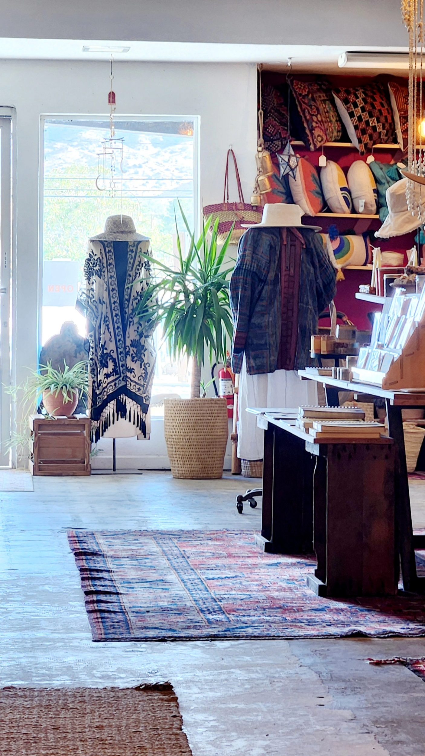 Interior of a boutique store with mannequins wearing bohemian-style clothing, assorted home decor items, and handmade crafts on display. Colorful pillows are stacked on a shelf, potted plants and rugs decorate the space. Sunlight streams through a window.